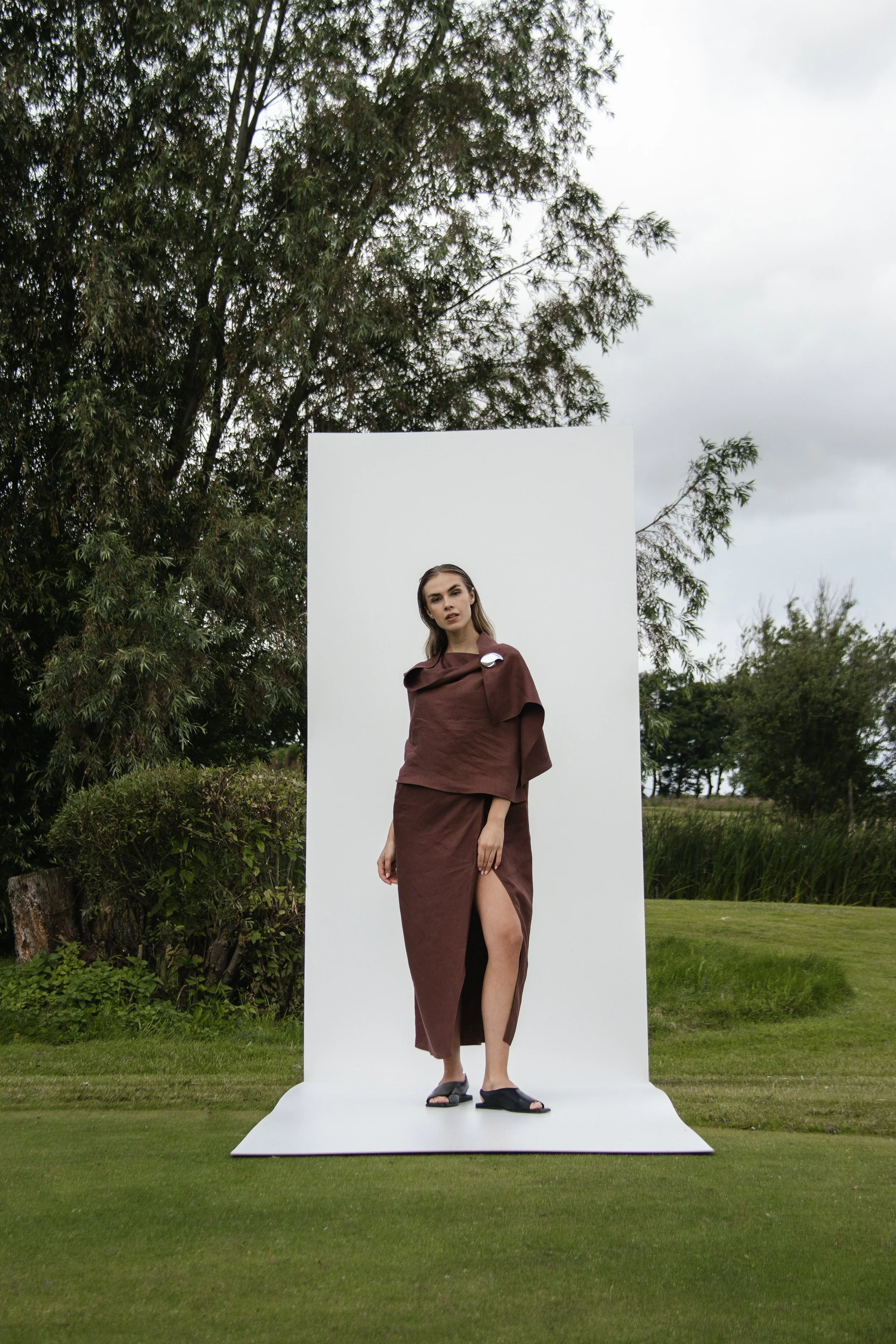 A woman wearing a brown dress with a slit standing on a white backdrop outdoors on grass with trees in the background.