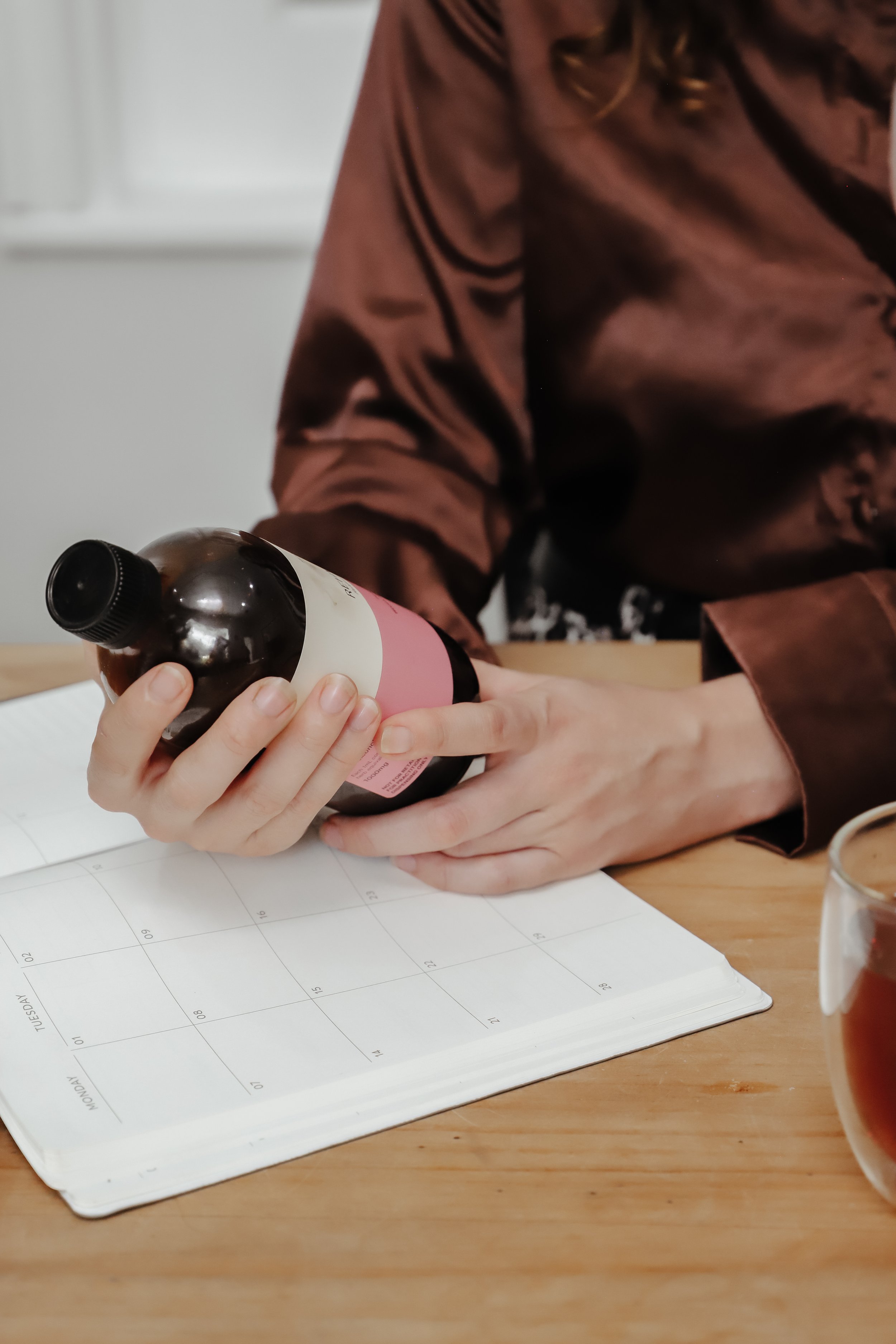 Person holding a brown bottle with a pink label over a calendar on a wooden table, with a glass of drink nearby.