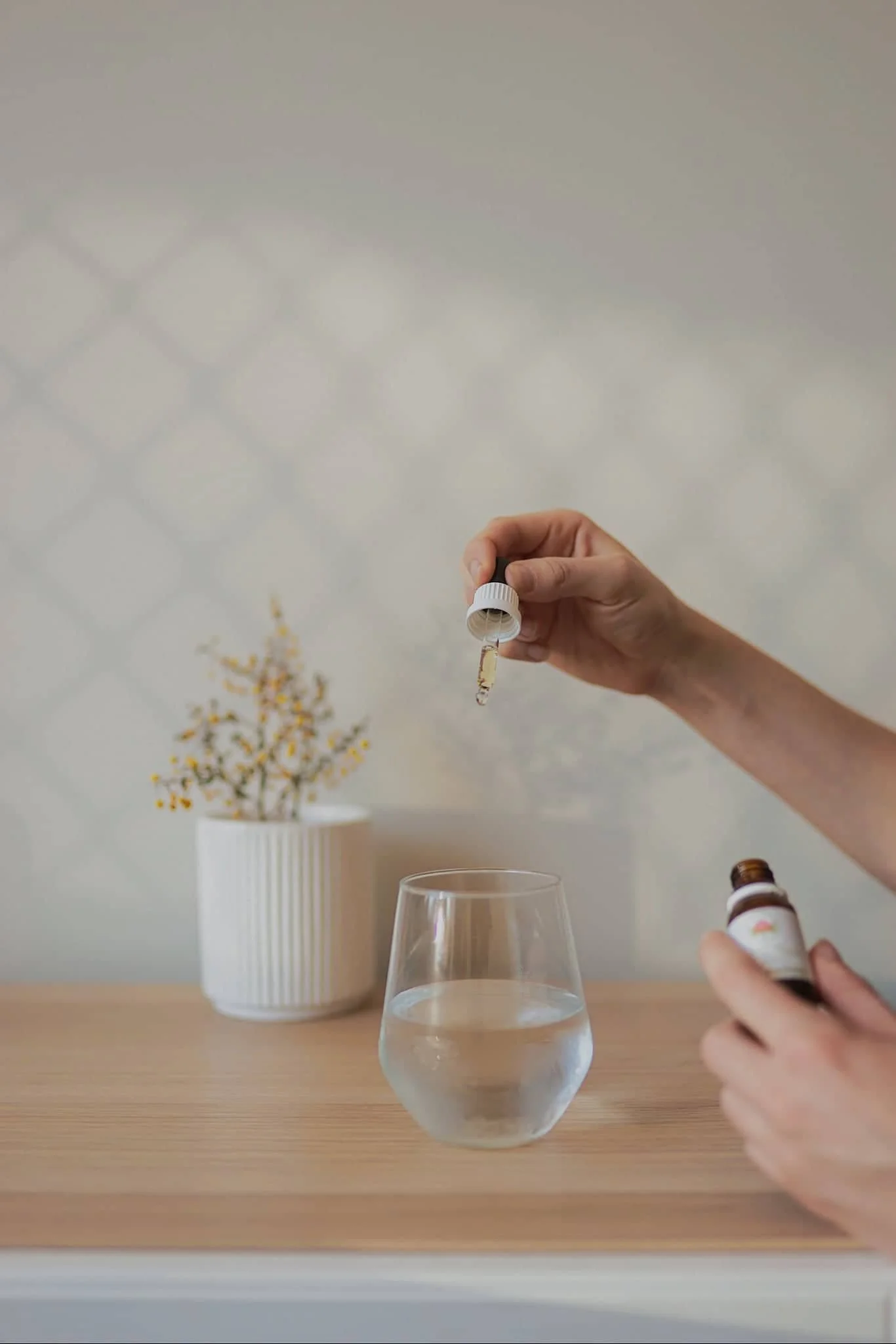 A person holding a dropper and a small bottle of liquid medicine or supplement over a glass of water on a wooden surface, with a white vase containing yellow flowers in the background.