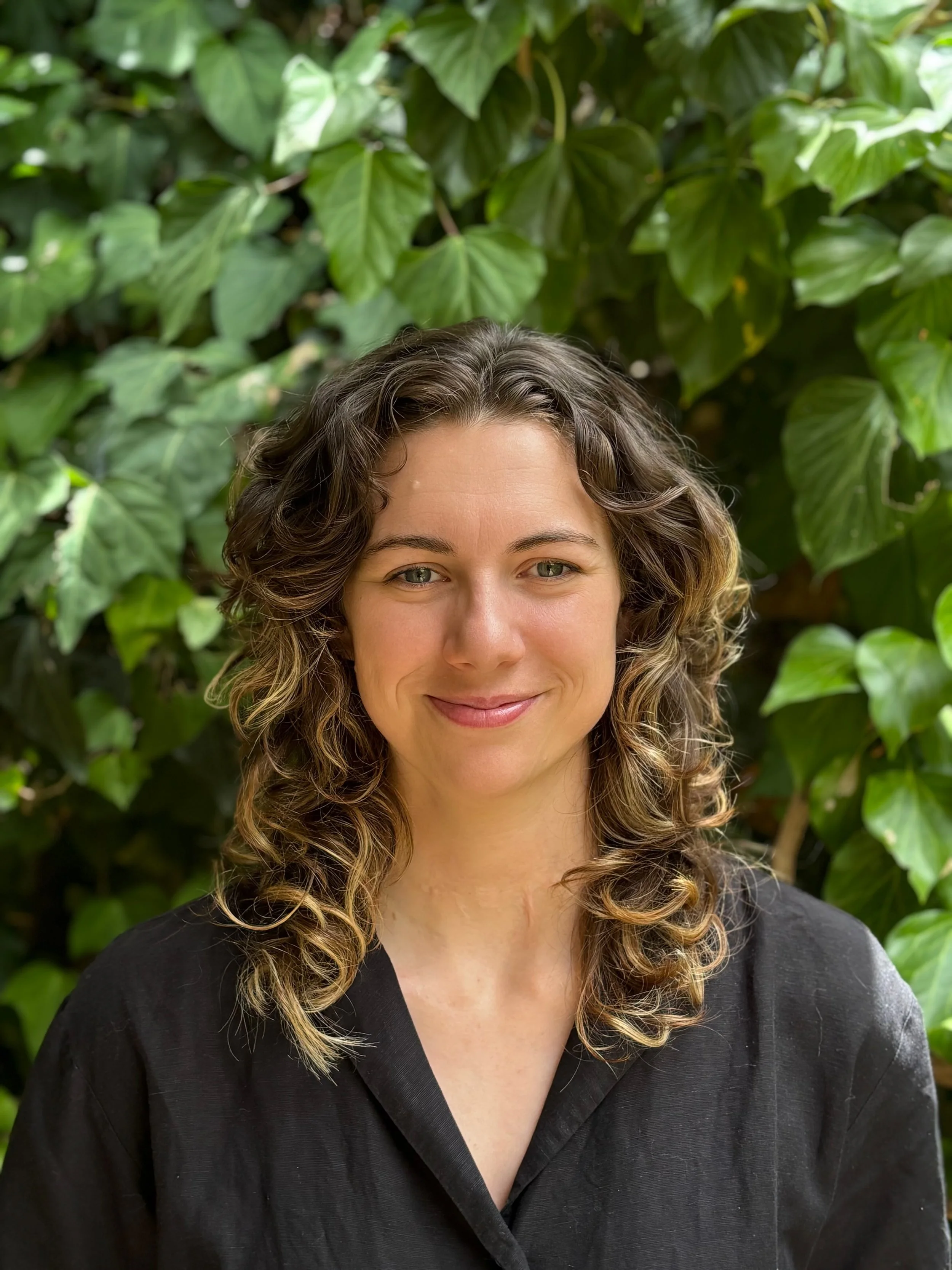 A woman with curly brown hair and a black top standing in front of green leafy plants, smiling at the camera.