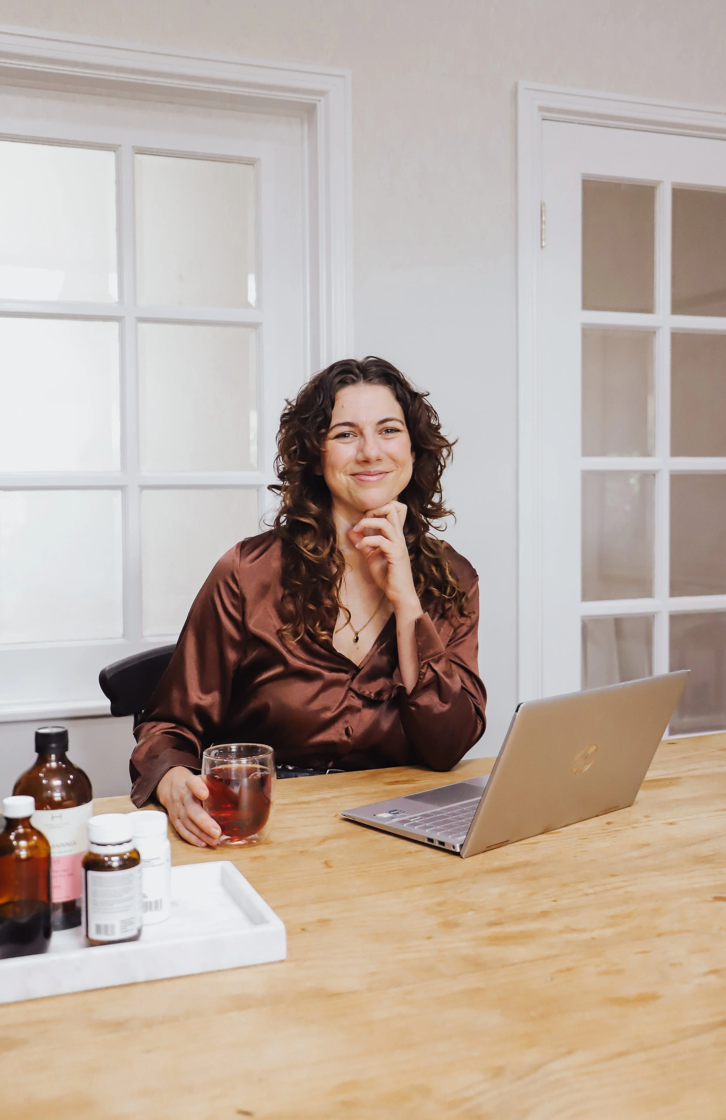 A woman with curly brown hair sitting at a wooden table, smiling and holding a glass of red beverage, with a laptop and medicine bottles on the table.