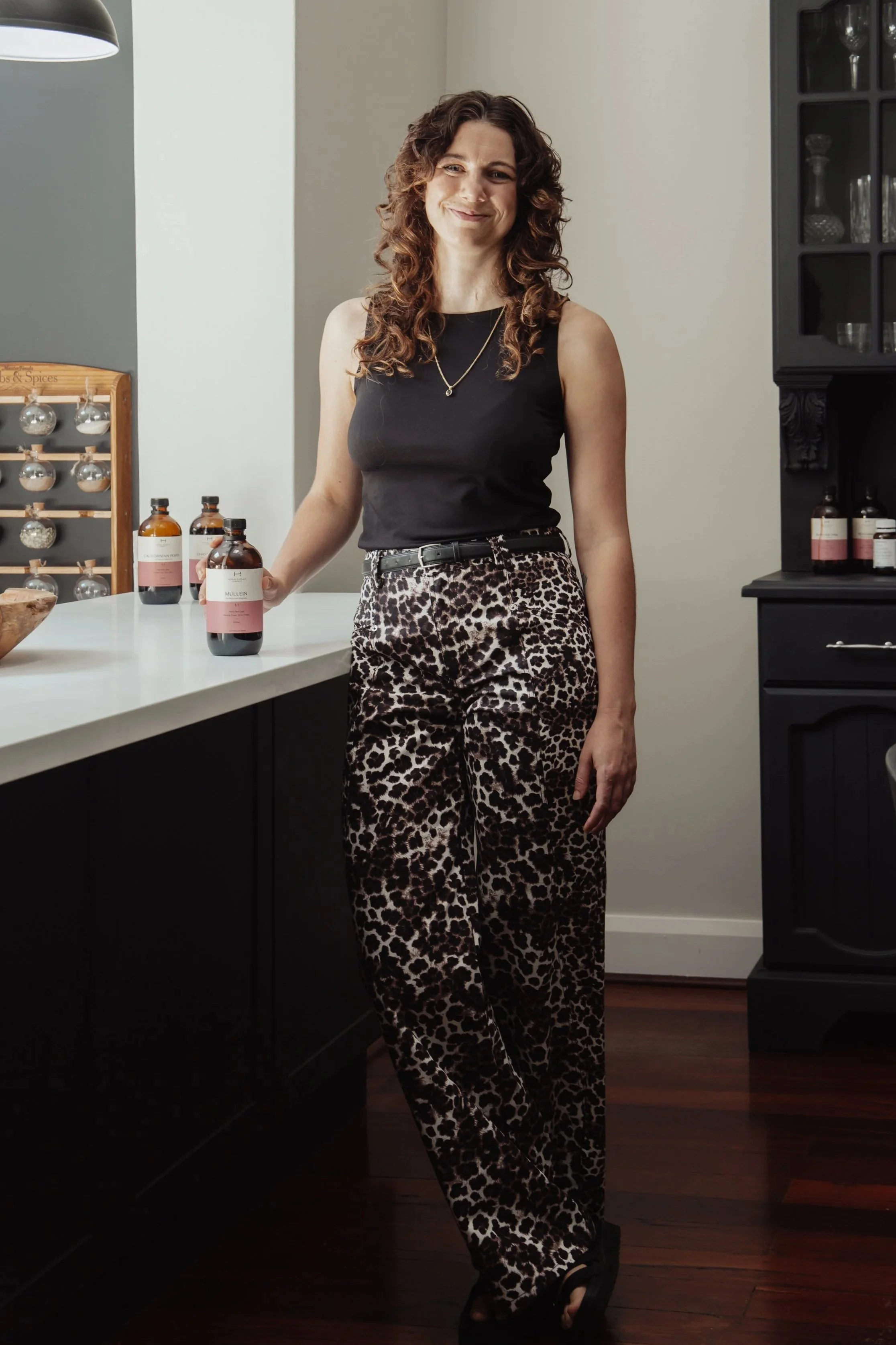 A woman with curly brown hair is standing in a modern kitchen, smiling, wearing a black sleeveless top and leopard print pants, with bottles of skincare or medication on the counter beside her.