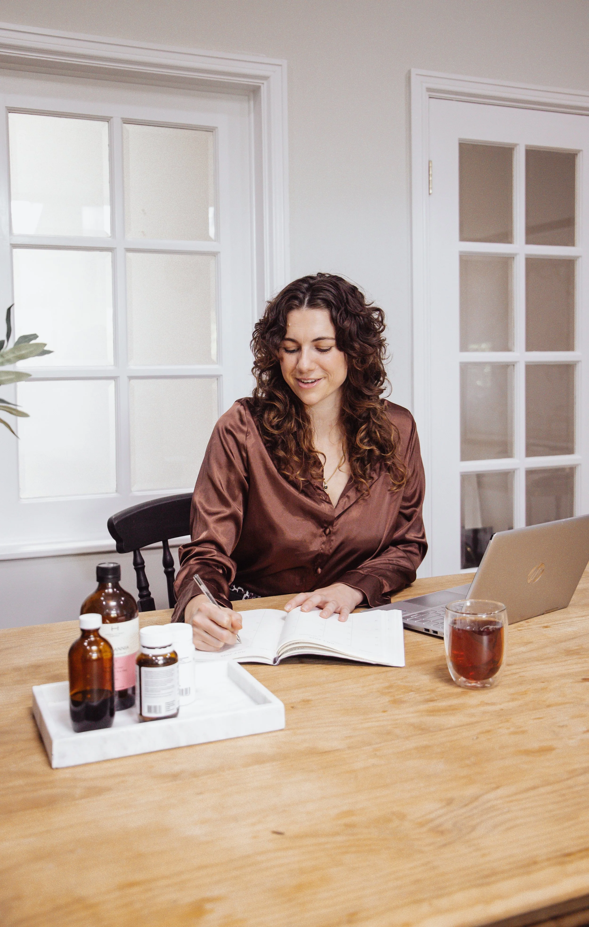 Woman sitting at a wooden table with bottles, a cup of tea, an open notebook, and a laptop, writing in her notebook inside a bright room with white walls and glass-paneled doors.