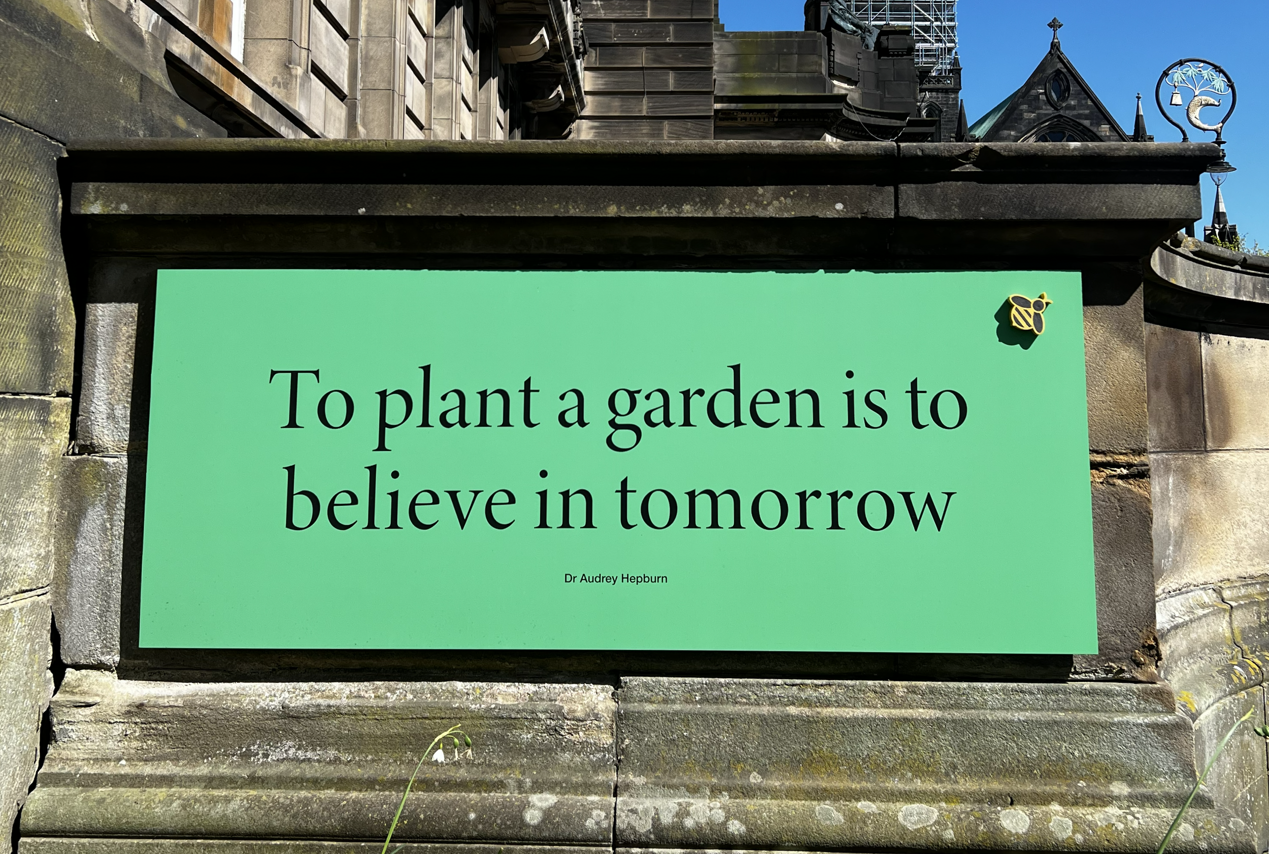 Yellow and black jesmonite bee on a green sign with quote by Dr Audrey Hepburn on wall of Glasgow Royal Infirmary medicinal garden