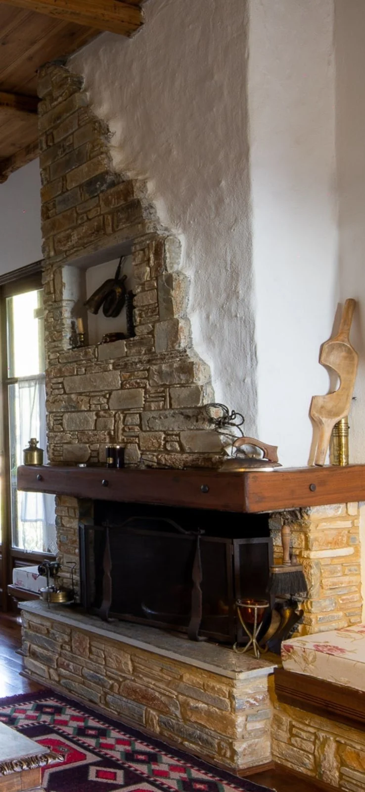 Living room corner with a stone fireplace, wooden mantel, and decorative objects, next to a window and patterned rug.