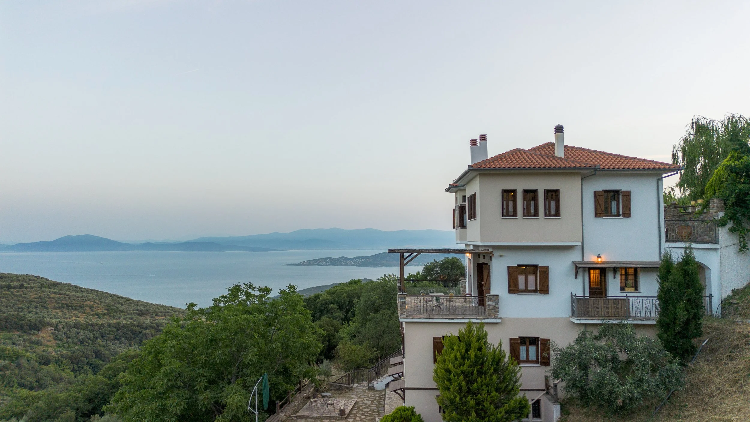 A white house with brown shutters and a red tiled roof sits on a hill overlooking a body of water with distant islands and mountains, surrounded by green trees.