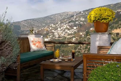 Outdoor patio with wooden furniture, yellow flowers in a pot, drinks, and a view of hillside houses in the background.