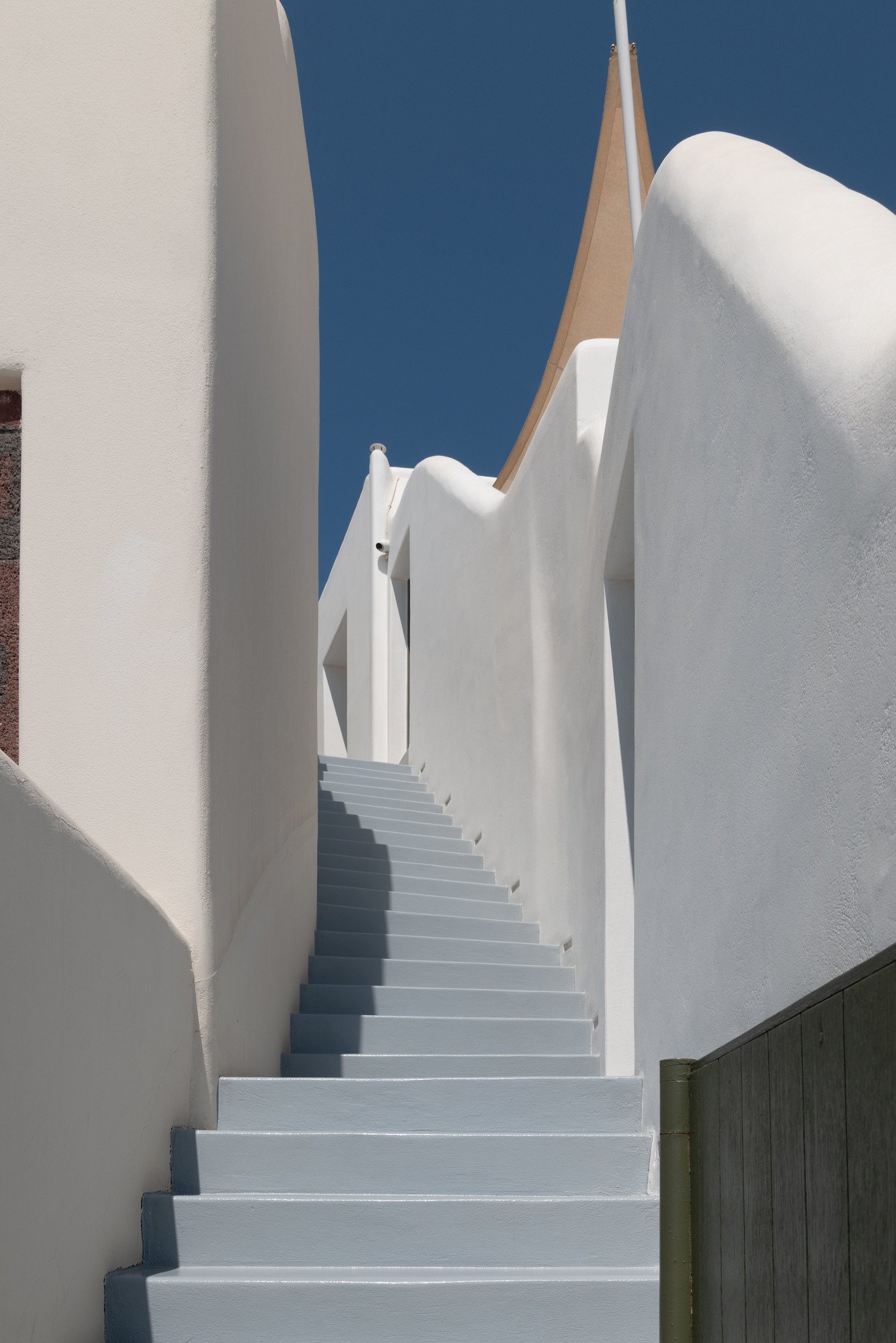 White staircase ascending between white walls towards a blue sky with a partial view of a beige and white structure at the top.