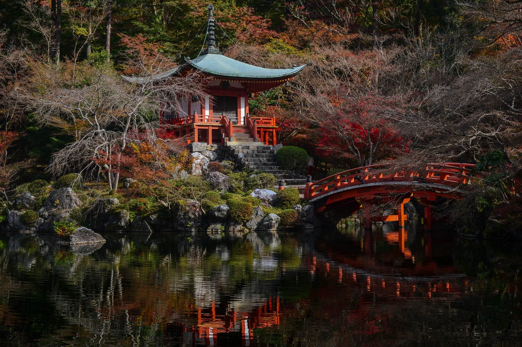 Daigoji Temple, home to Kyoto’s Oldest Pagoda and Gardens - A traditional Japanese pagoda-style structure on a small hill, surrounded by trees with autumn foliage, next to a pond with reflections of the structure and trees.