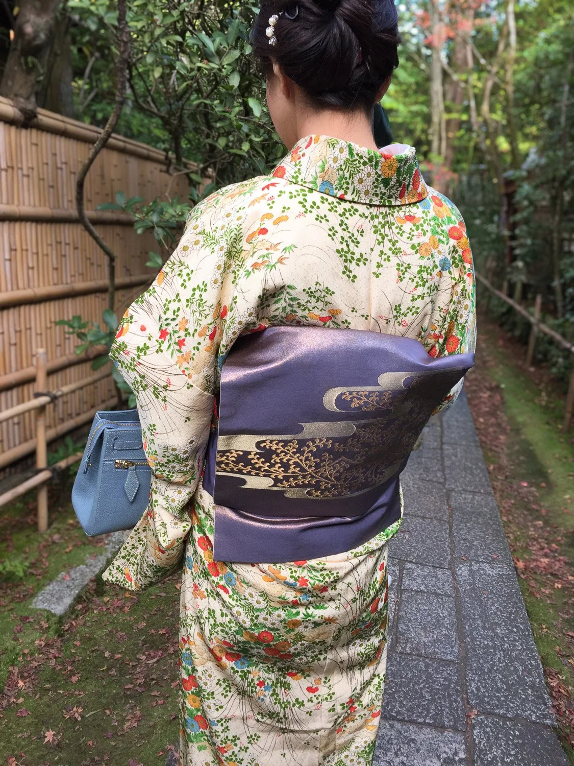 A woman dressed in a traditional Japanese kimono with floral patterns, standing on a stone pathway in a garden with wooden fencing and green foliage, holding a light blue handbag.