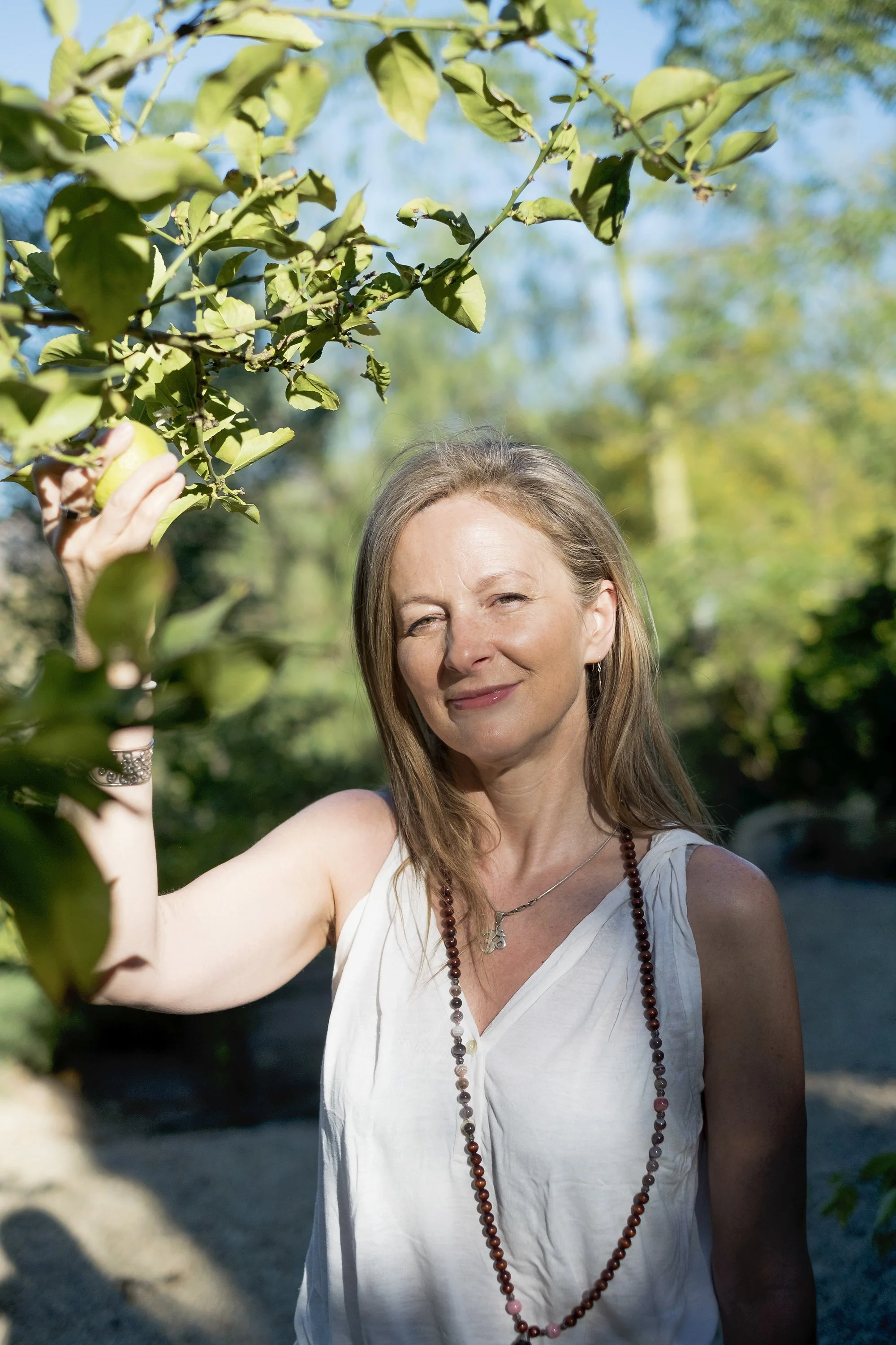 A woman with long light brown hair, wearing a white sleeveless blouse and a beaded necklace, stands outdoors in sunlight, smiling gently while holding onto a branch of green leaves from a tree.