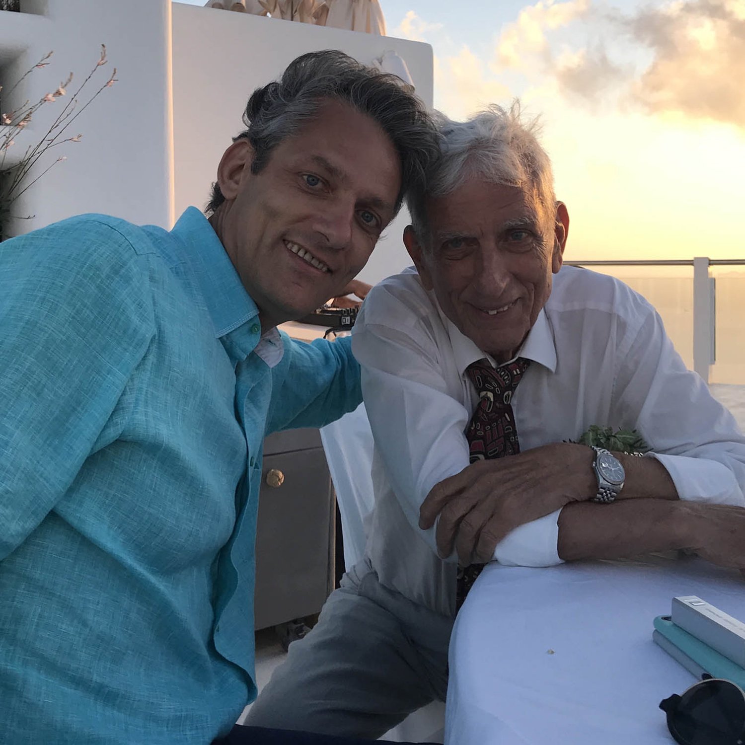 A modern photograph of two men sitting together at an outdoor table, smiling warmly at the camera. The younger man, dressed in a turquoise shirt, leans affectionately toward the older man, who is wearing a white shirt and patterned tie. The background features a serene sunset over the ocean, evoking a sense of connection and shared memories.