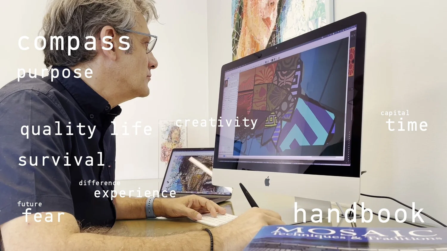 Designer at an iMac with a stylus, a mosaic book on the desk and a geometric artwork on screen