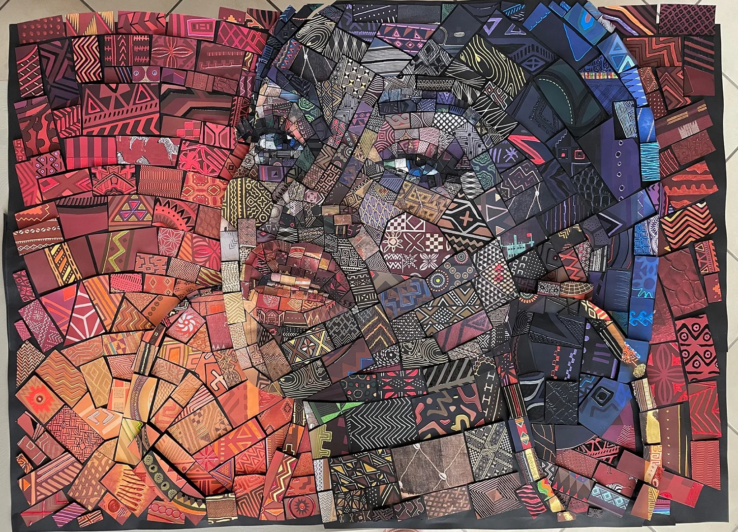 Handmade paper mosaic of a face with blue eyes in orange, purple and black patterned cut paper tiles, lying flat
