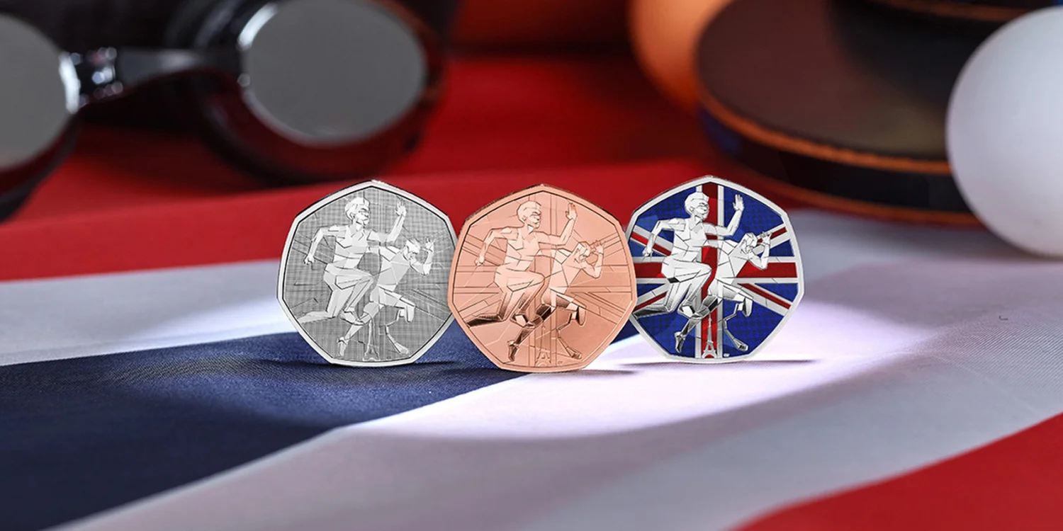 Three commemorative coins displayed prominently on a surface adorned with the Union Jack flag. Each coin features a unique color scheme and metallic finish, showcasing different aspects of the Olympic and Paralympic spirit. The left coin is silver, depicting two athletes—one standing and one seated—in a dynamic pose. The middle coin is rose gold, highlighting an athlete in motion, symbolizing determination and effort. The right coin is blue with white accents, featuring the same dual-athlete design, emphasizing unity and collaboration. The coins are arranged neatly, with headphones and other objects blurred in the background, drawing attention to their intricate details and symbolic imagery.