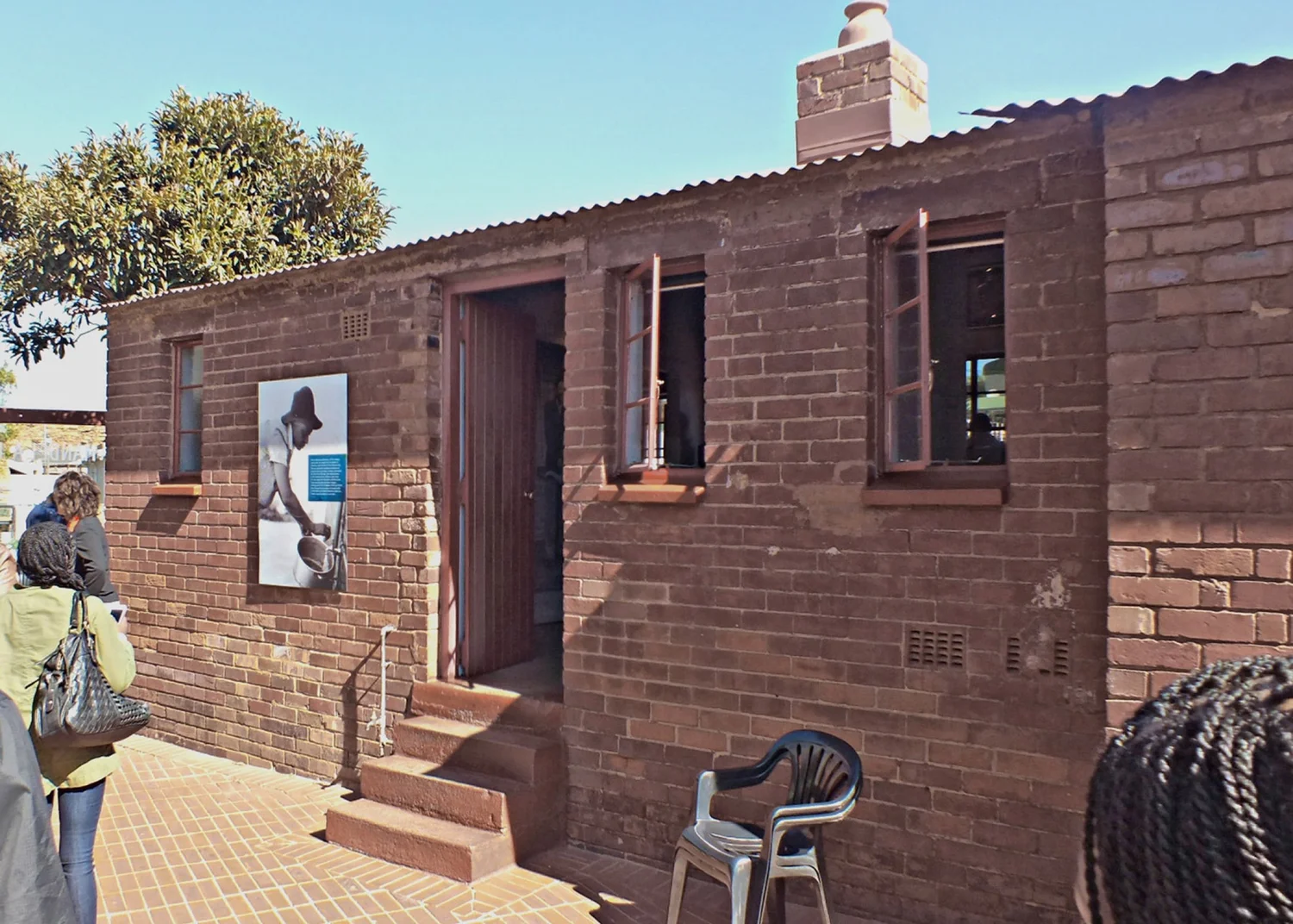 Small brick house with a corrugated roof and open door, a framed photo mounted on its wall, visitors standing in the sunshine