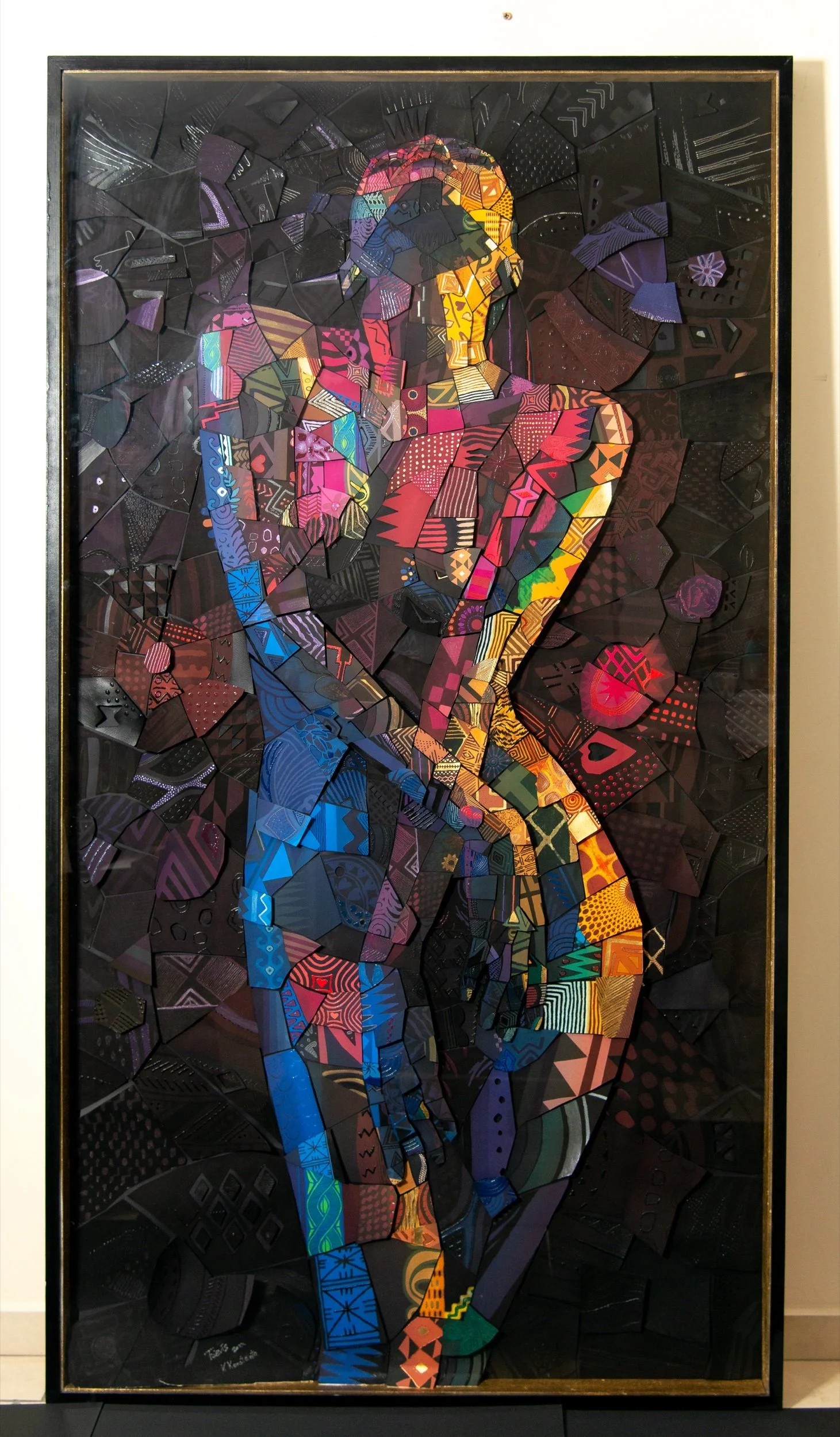 Tall framed mosaic of a standing figure made of colorful patterned paper tiles that glow against a dark black background