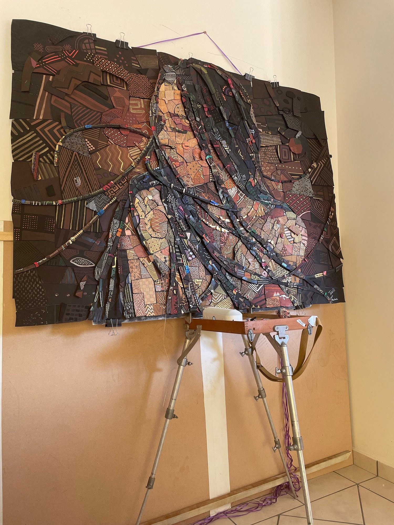 Large handmade paper mosaic of a woman with long braids, pinned to a wall above an easel, built from dark patterned tiles