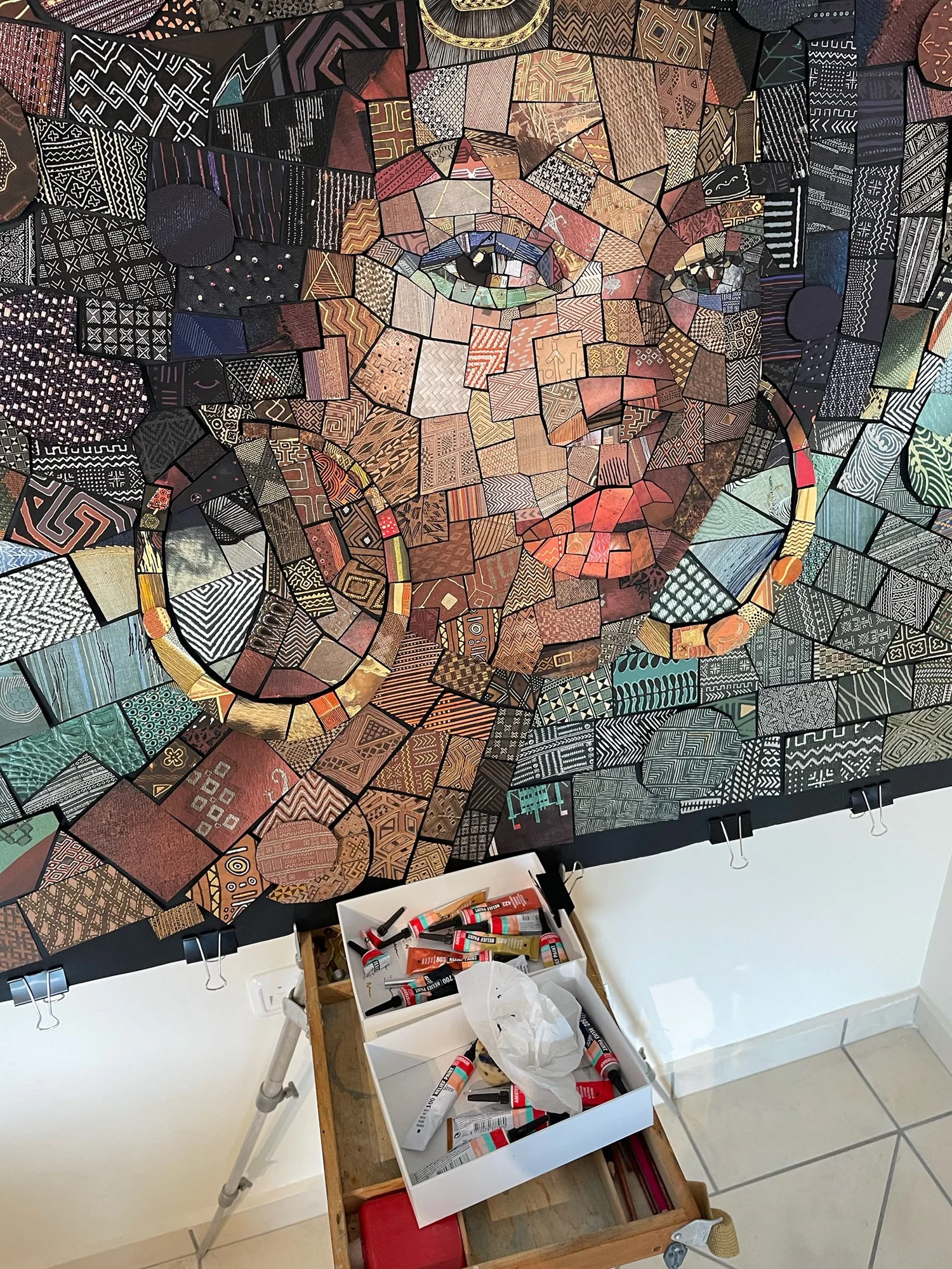 Close detail of a handmade paper mosaic with an eye, hoop earring and patterned tiles, with a tray of glue tubes below