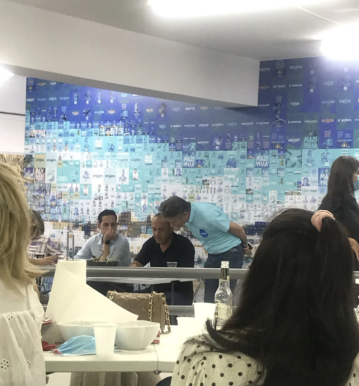 A group of people are gathered around a table during an event, with a large mosaic mural in the background. The mural displays a collage of images and text, adding a vibrant backdrop to the scene.