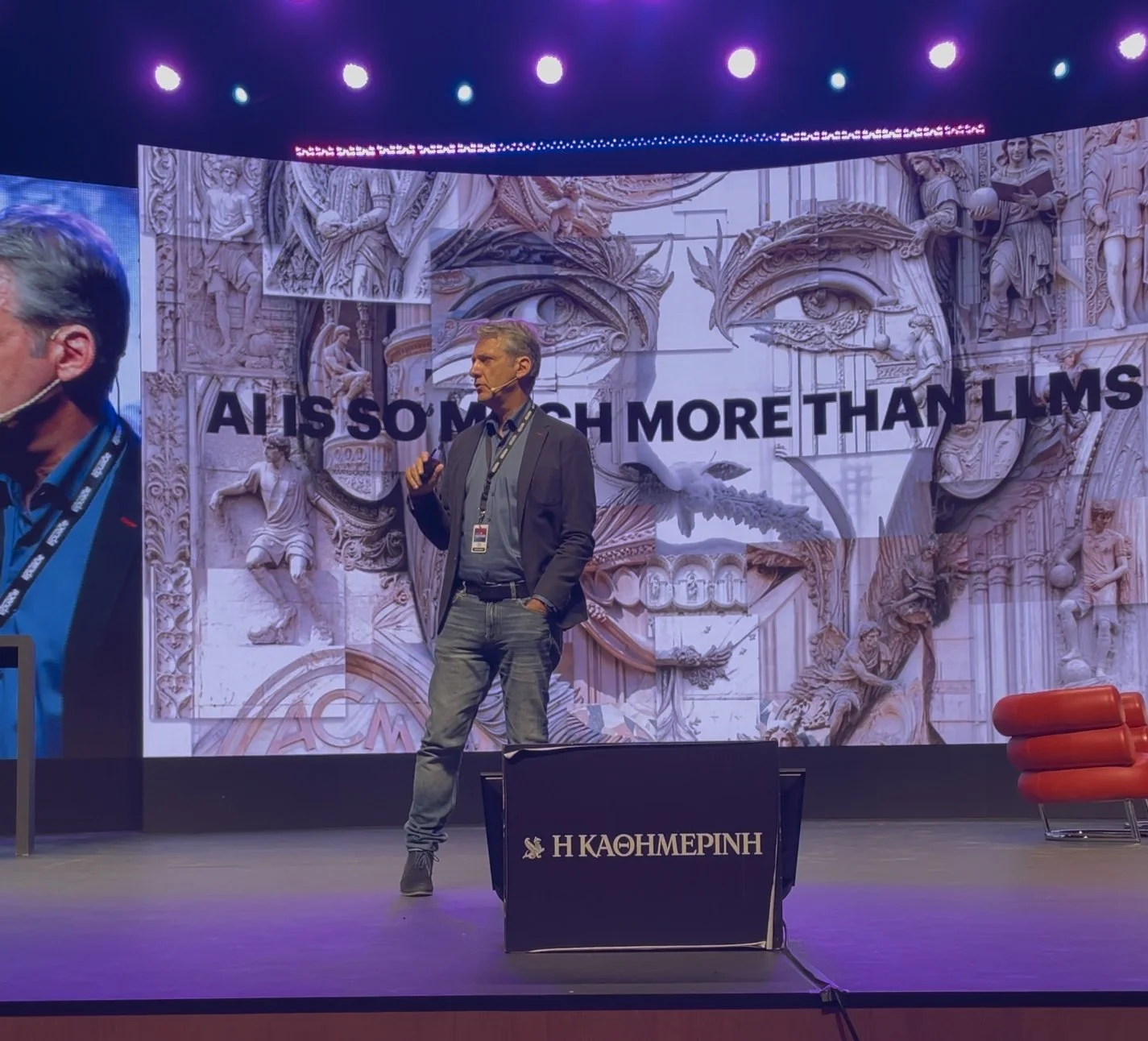 Charis Tsevis on stage holding a microphone, in front of a screen showing a mosaic of a classical face and sculpture fragments with the words “AI is so much more than LLMs”.
