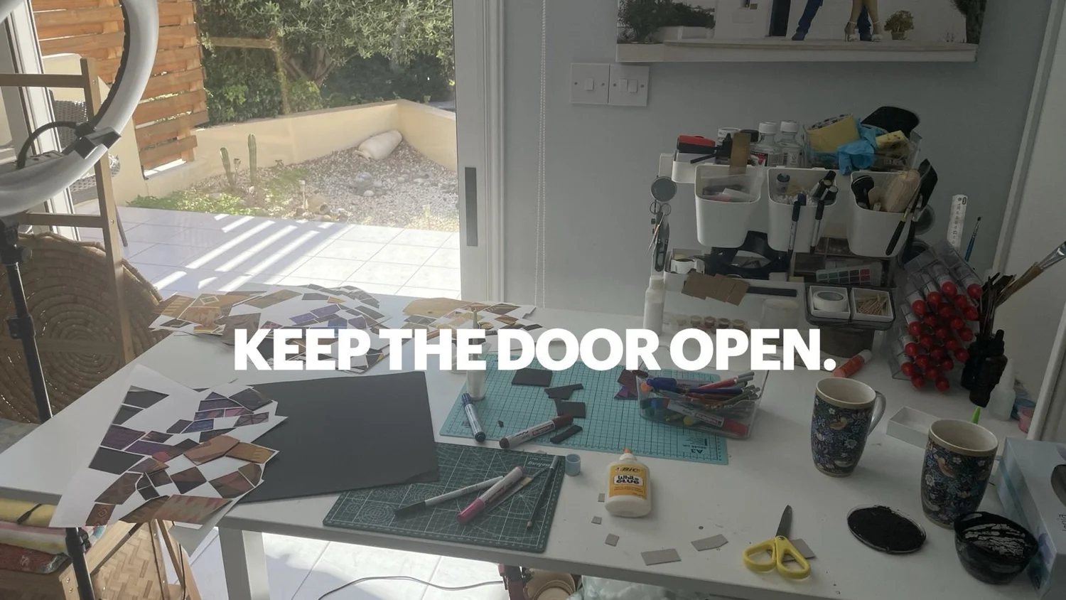 A sunlit studio table covered with cut paper tiles, cutting mats, glue, markers and scissors, in front of an open door to a terrace, with the words “Keep the door open.”