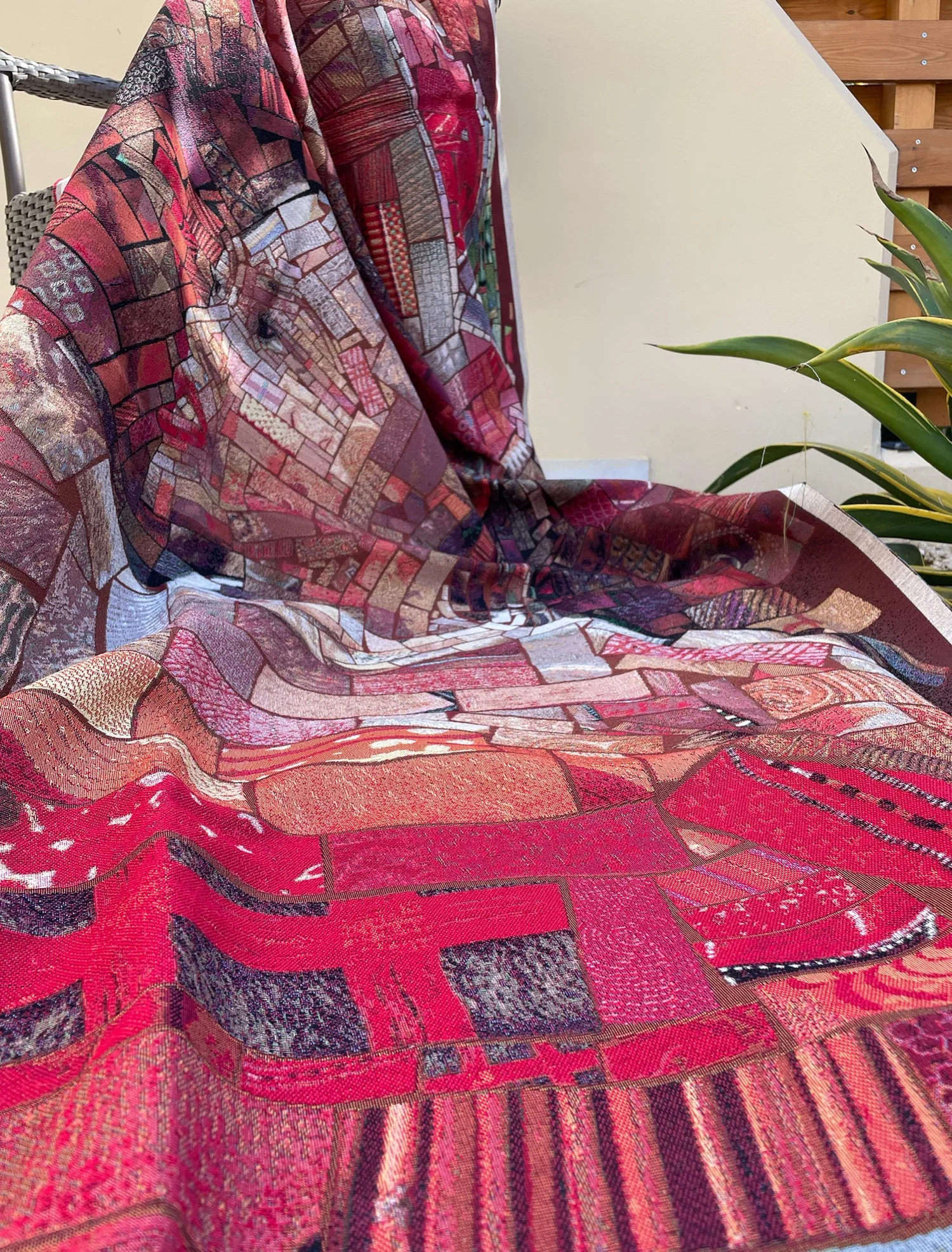 Red woven mosaic fabric draped over a chair outdoors, with a face visible in the folds and patterned brick-like tiles