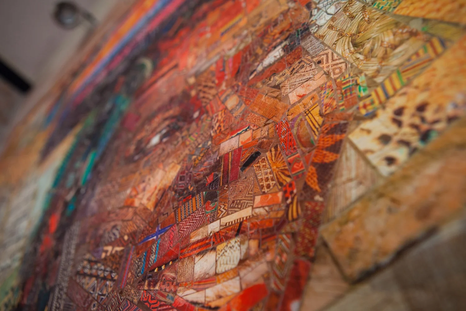 Tilted close-up of a large mosaic print on a wall, showing a face built from small orange and red patterned tile pieces
