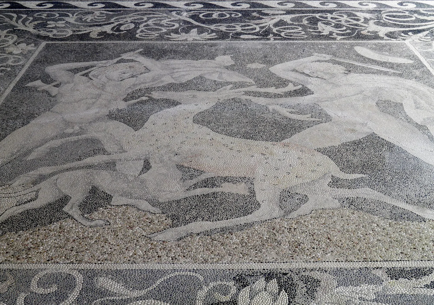 Greek pebble mosaic of two hunters and a dog attacking a stag, in pale stones on a dark ground