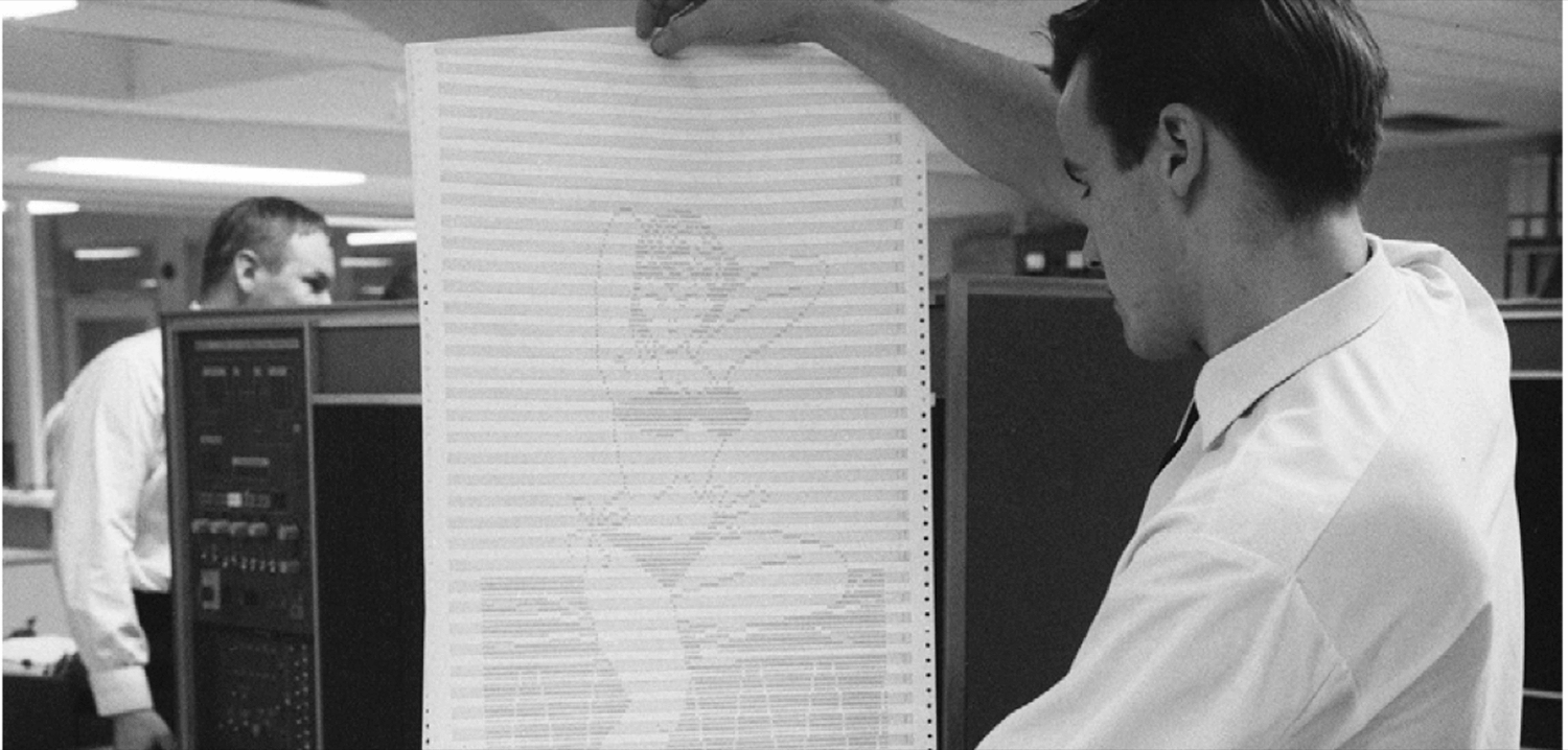 A black-and-white photograph showing a man standing in a workspace, holding a tall sheet of paper covered in dense printed text. Another person is partially visible in the background, focused on work near a piece of electronic equipment. The scene suggests a historical context of early computing or data processing.