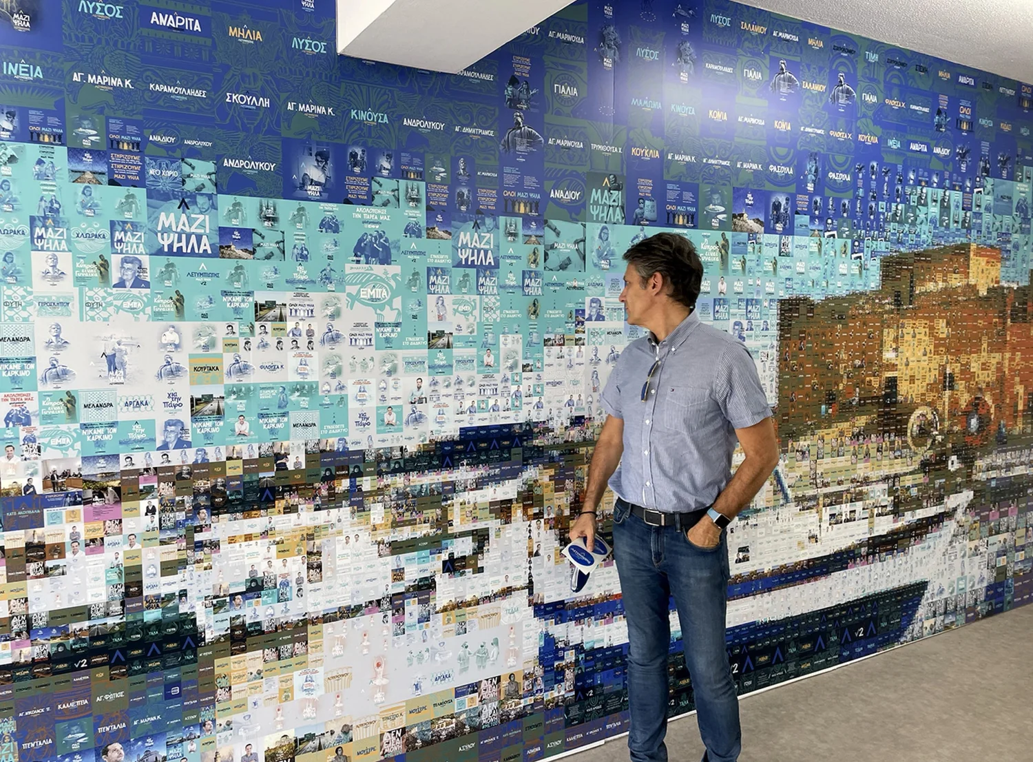 Charis Tsevis stands in front of a vibrant mosaic mural, which is composed of numerous small images and text tiles. The mural features a blend of colors and patterns, creating a dynamic visual effect.