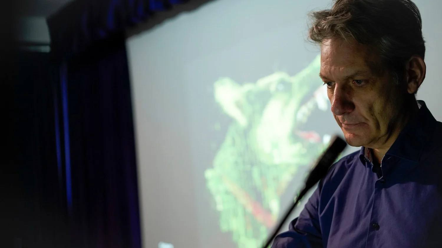 Charis Tsevis in profile beside a microphone, a green mosaic artwork projected behind him