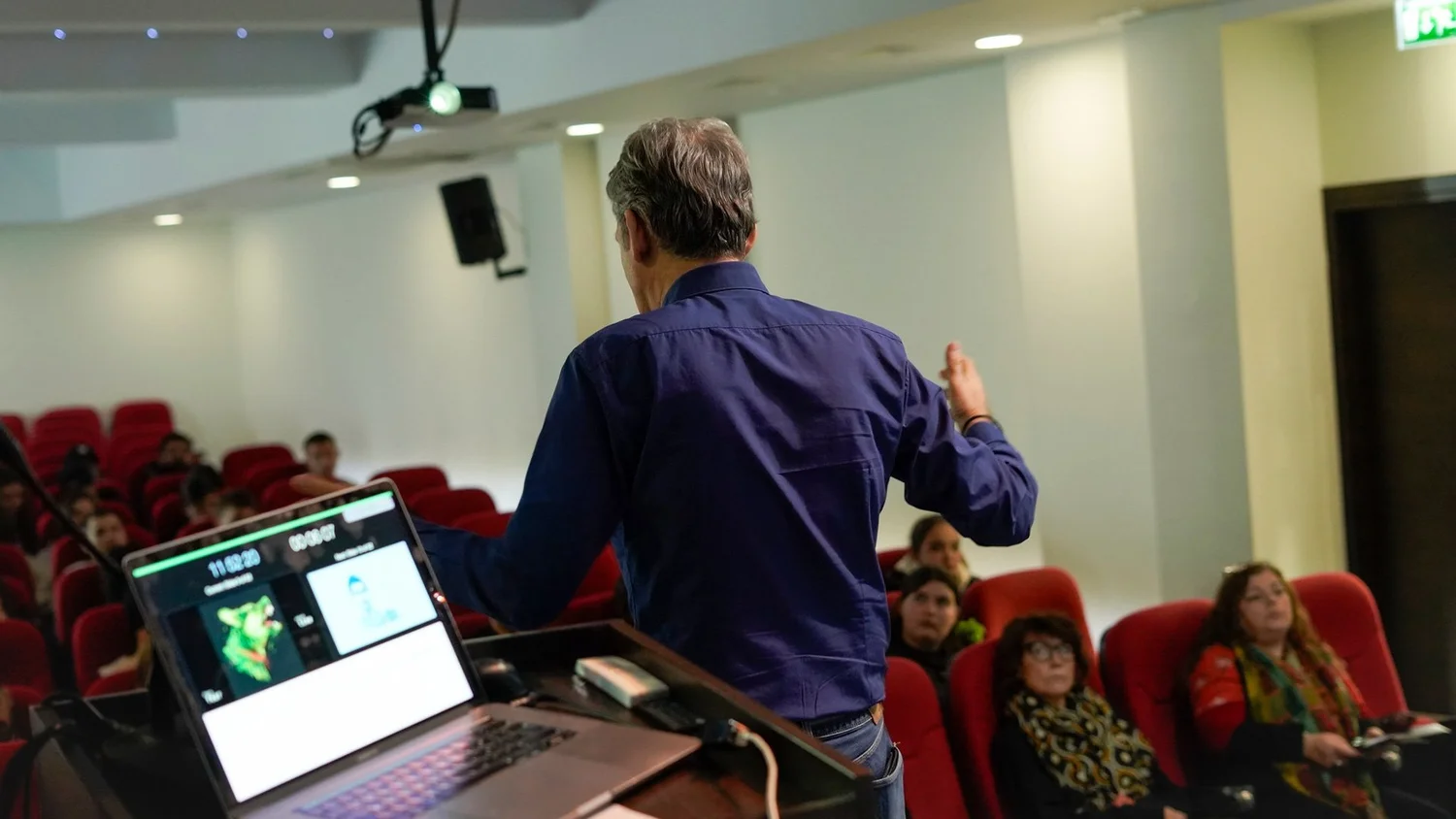 Charis Tsevis seen from behind, gesturing toward students in red seats, a laptop showing slides