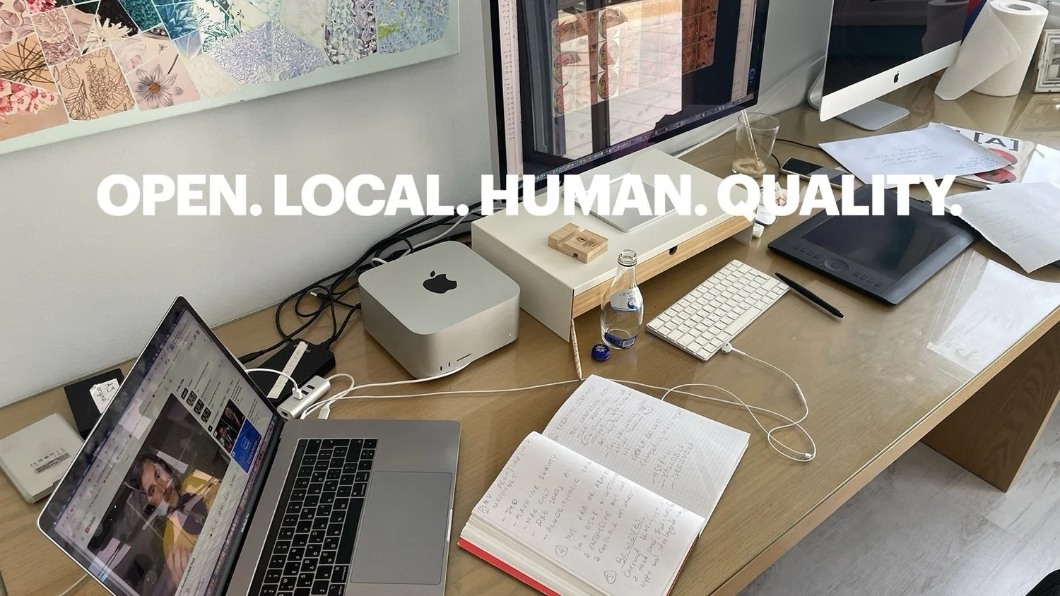 A desk with a laptop, a small desktop computer, a drawing tablet and an open handwritten notebook, with the words “Open. Local. Human. Quality.”