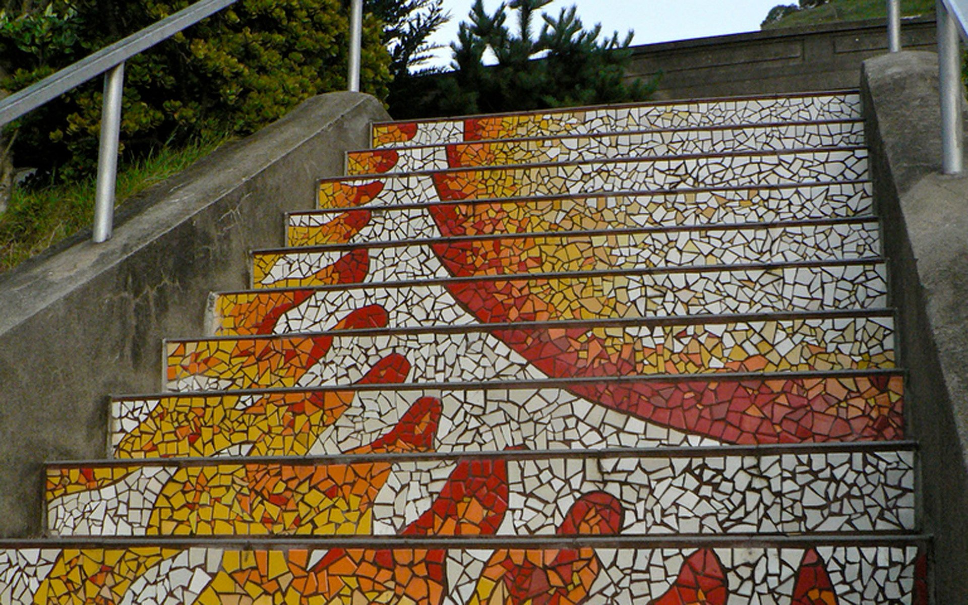 Detail of the 16th Avenue Tiled Steps with bright orange and red mosaic tiles forming a stylized sunburst pattern, radiating warmth and energy at the base of the staircase.
