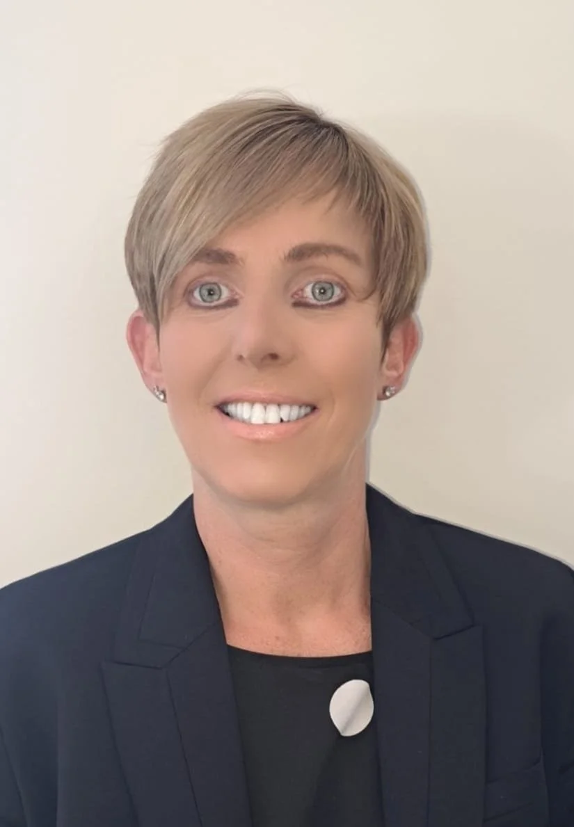 A man with dark curly hair, glasses, and a beard, wearing a dark green sweater and looking to the side, indoors with a blurred plant in the background.