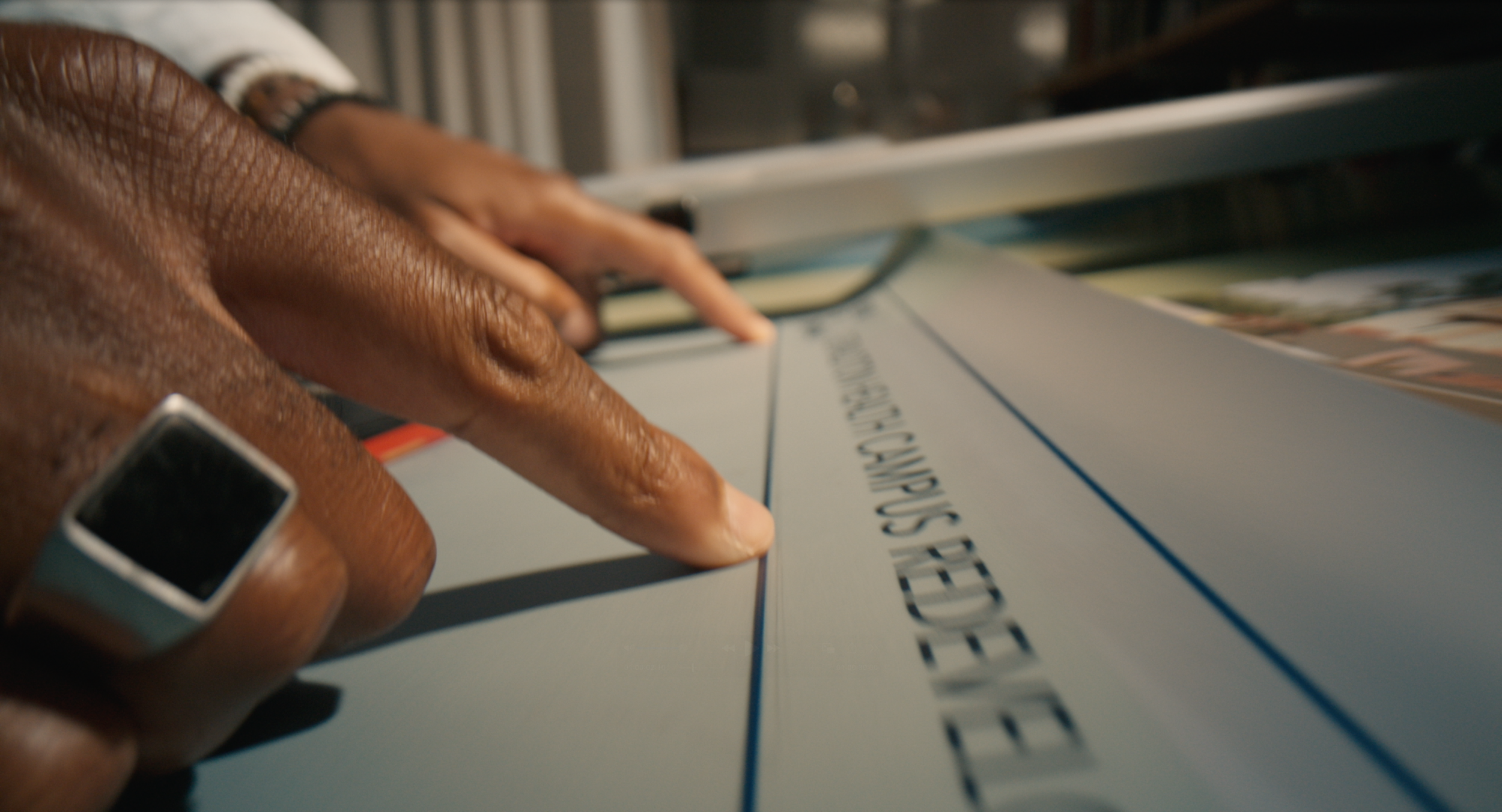 Close-up of a person's hand with dark skin and a smartwatch, touching or pointing at a sheet of paper or card with printed text, possibly at a library or office.