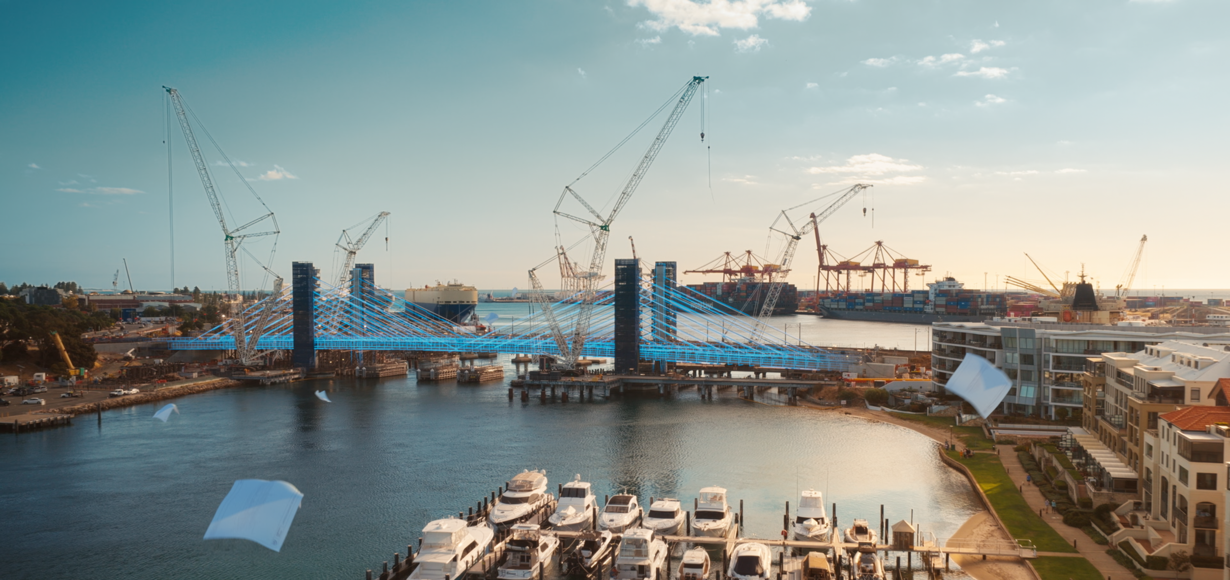 A harbor with yachts docked in the foreground and a bridge under construction with multiple cranes in the background, illuminated by sunlight.