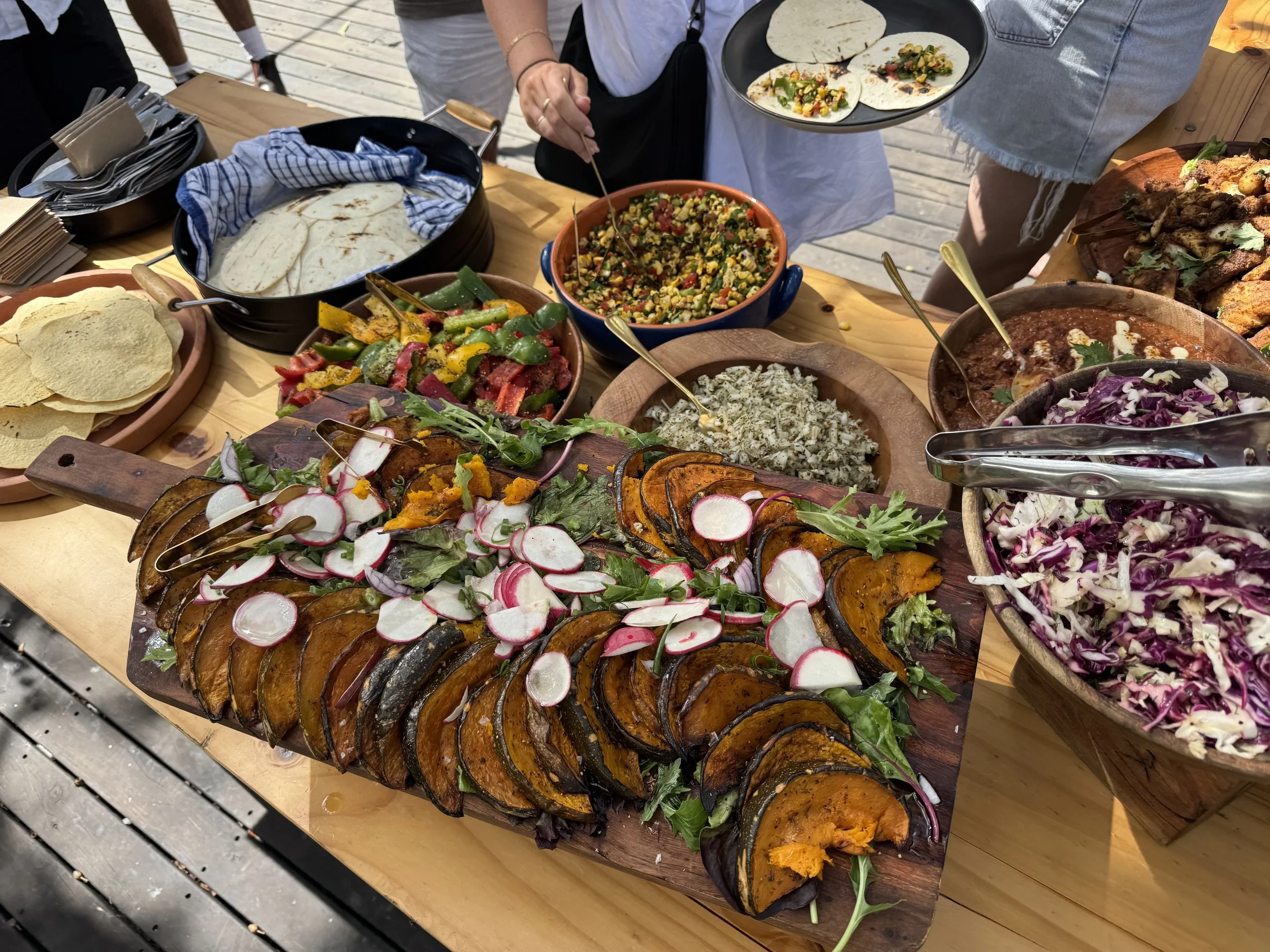 Outdoor table with various Mexican dishes including grilled vegetables, guacamole, salsa, tortillas, and salad, with people serving themselves.