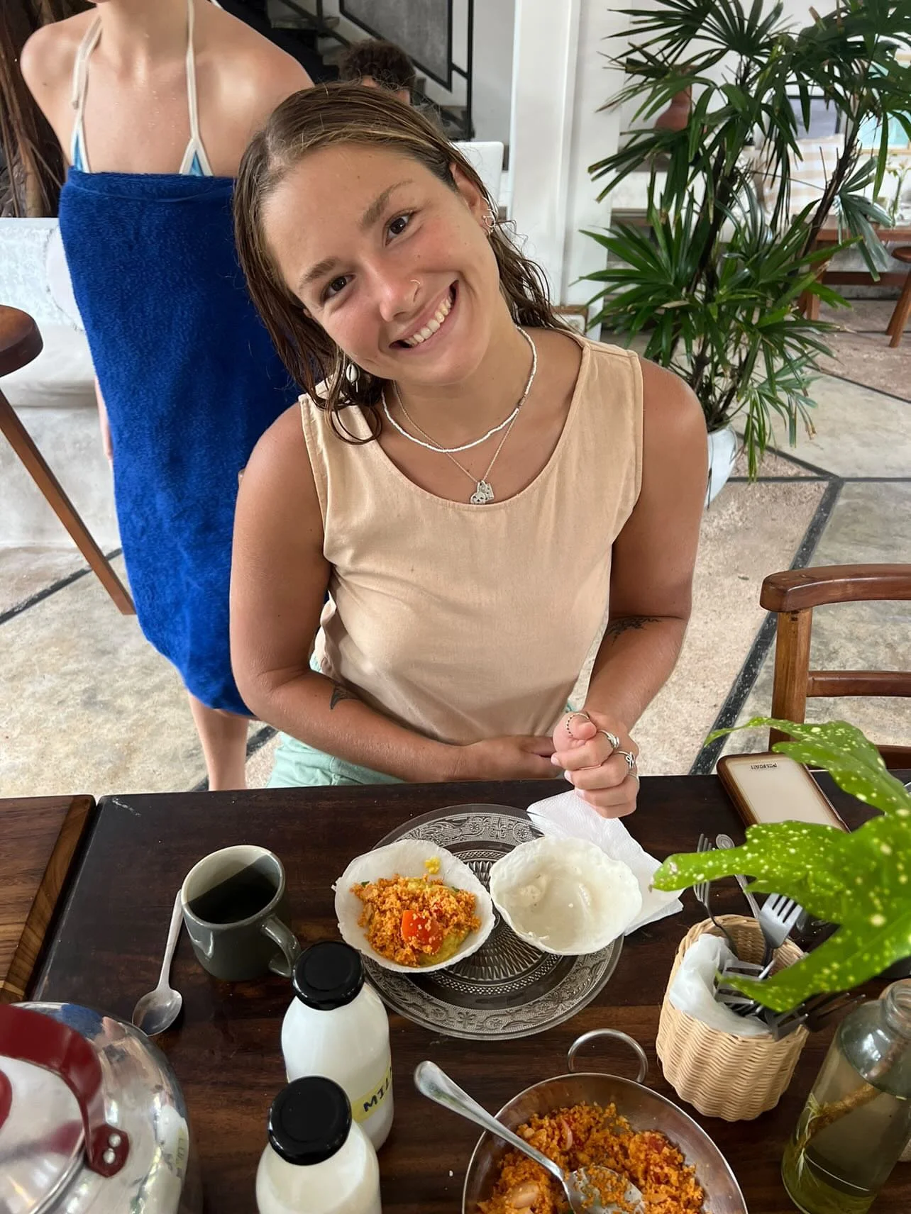 A woman with a big smile sitting at a dining table with food, including rice and a curry dish, in front of her. Behind her is another person with a towel on their shoulders. There are plants and dining furniture in the background.