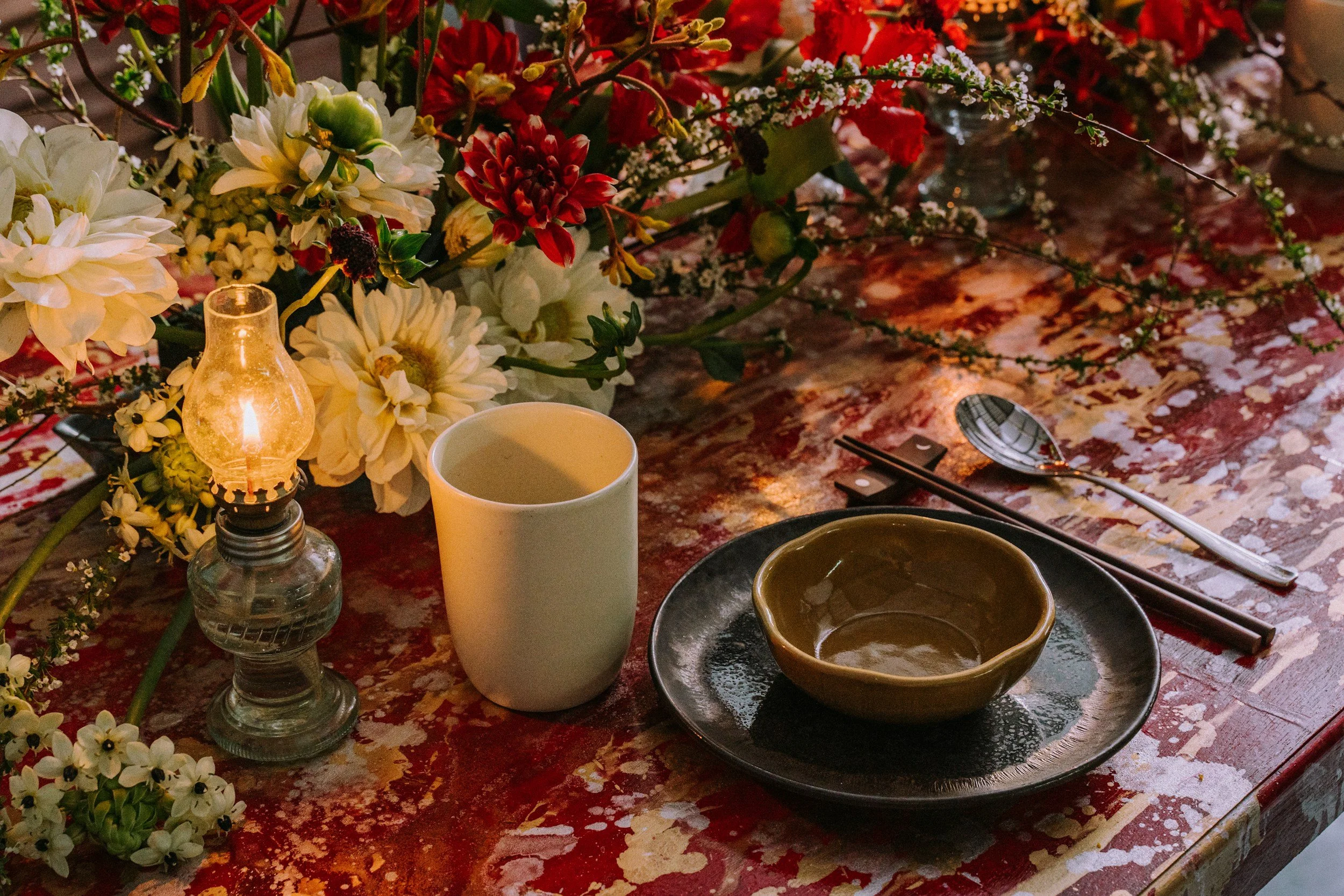 A table decorated with a floral arrangement of white and red flowers, a lit candle in a glass holder, a white mug, and an empty brown bowl on a red patterned tablecloth. Silverware is placed beside the bowls.