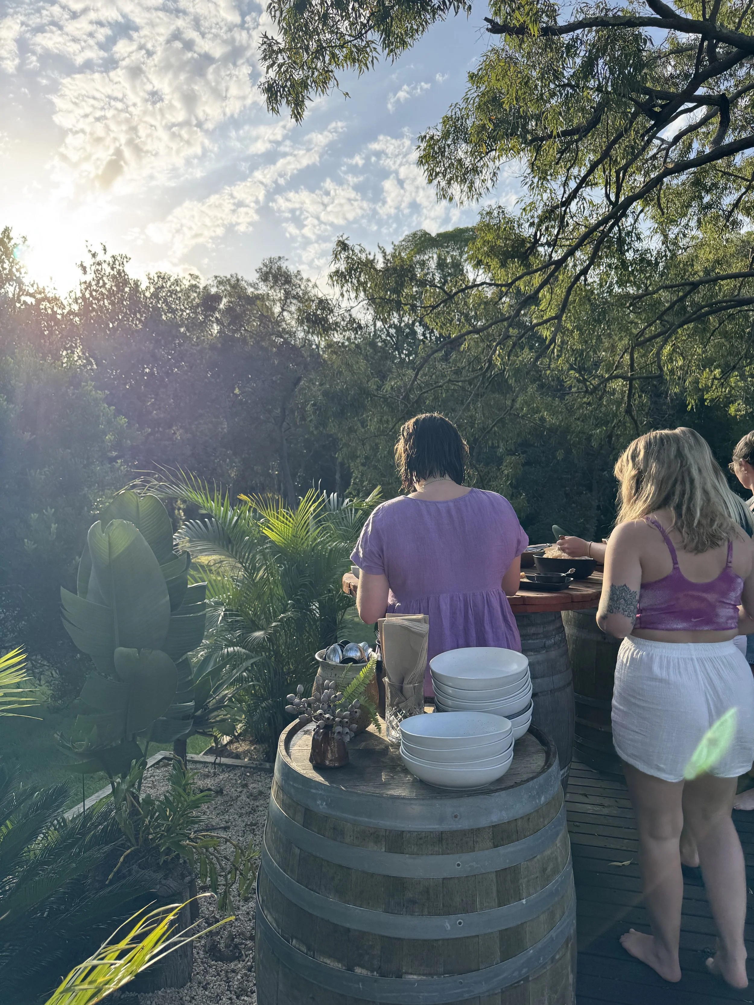 People gathered outdoors near a table with bowls and utensils, surrounded by lush greenery and trees, during a sunny day with partly cloudy sky.