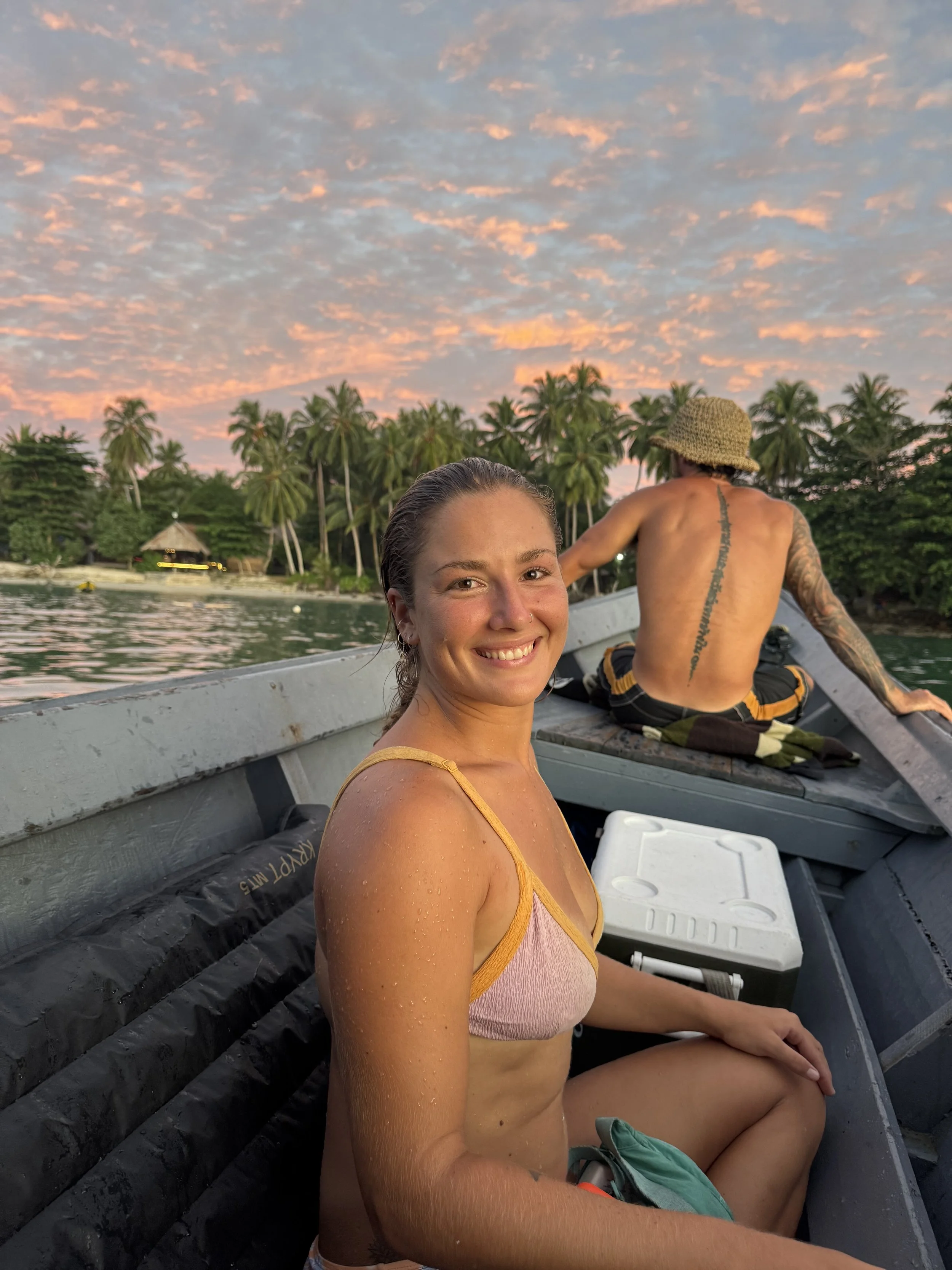 Smiling woman in a boat with a tropical shoreline and palm trees at sunset, with a tattooed man in a straw hat sitting behind her.