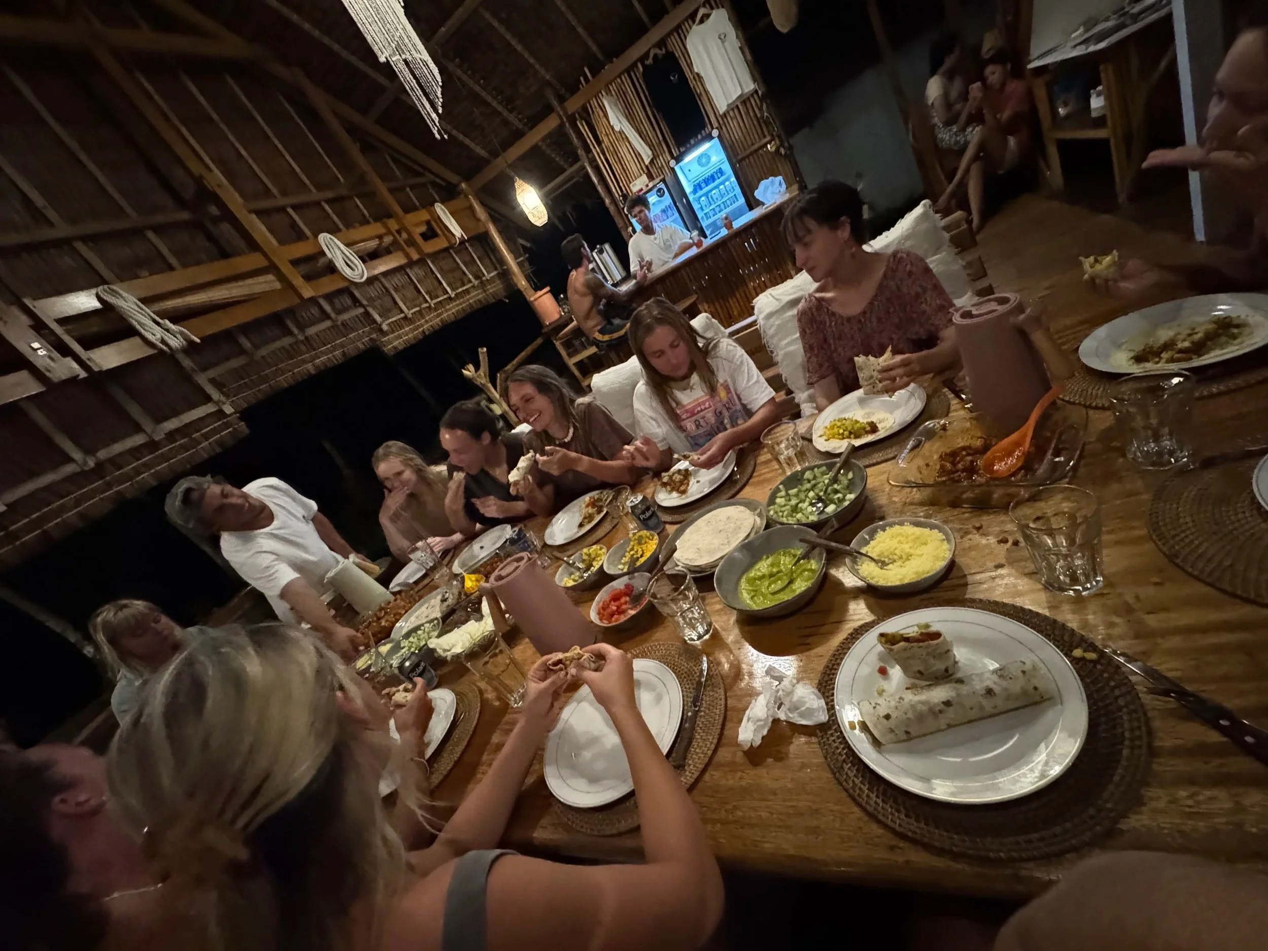 People sitting around a long wooden table enjoying a meal in a rustic, open-air setting with wooden beams and woven decor. There are plates of food, bowls of salad, and drinks on the table, and a bar in the background.