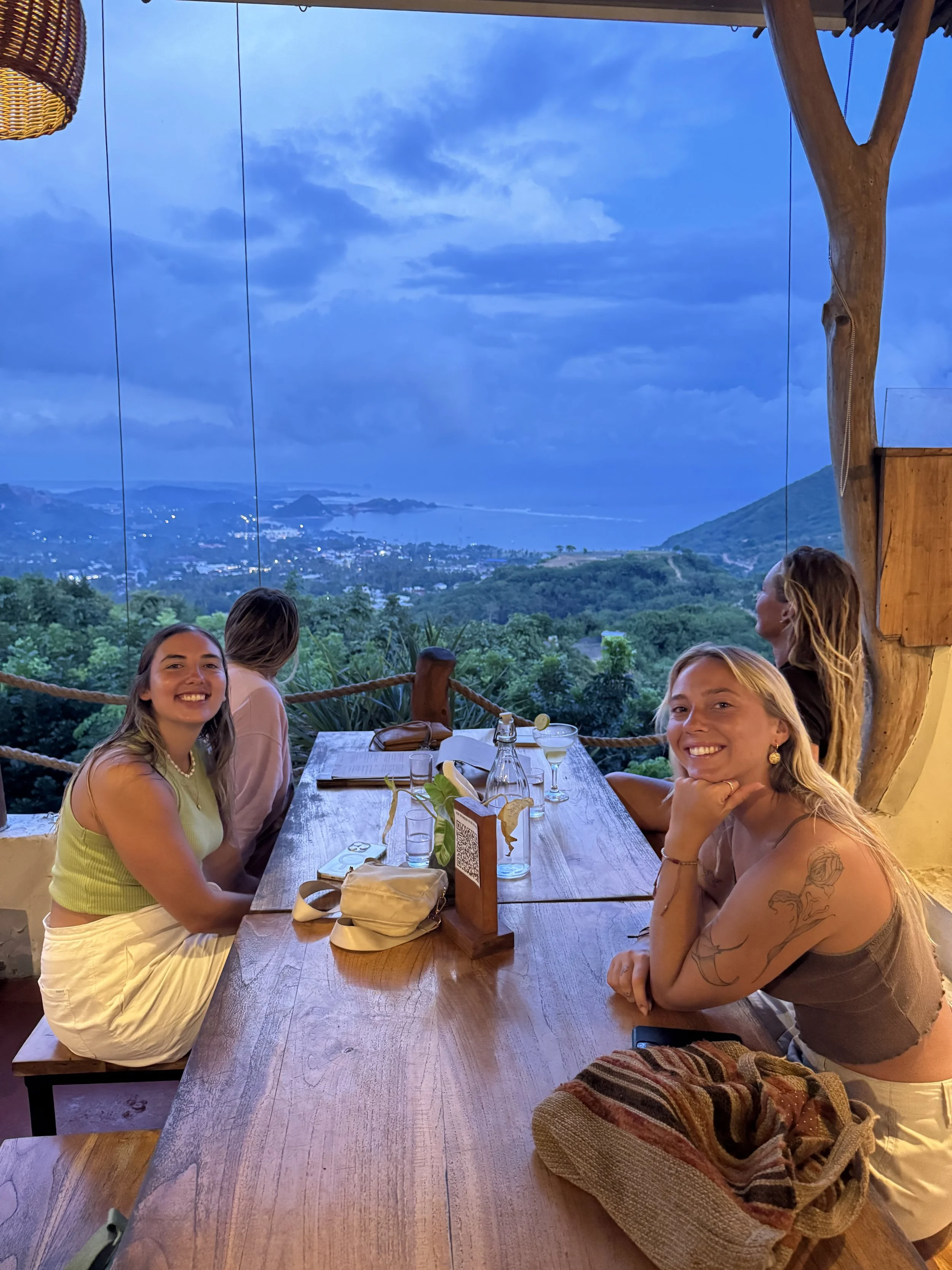 Four women sitting at a wooden dining table on an outdoor patio, enjoying a scenic view of mountains and a body of water during twilight.