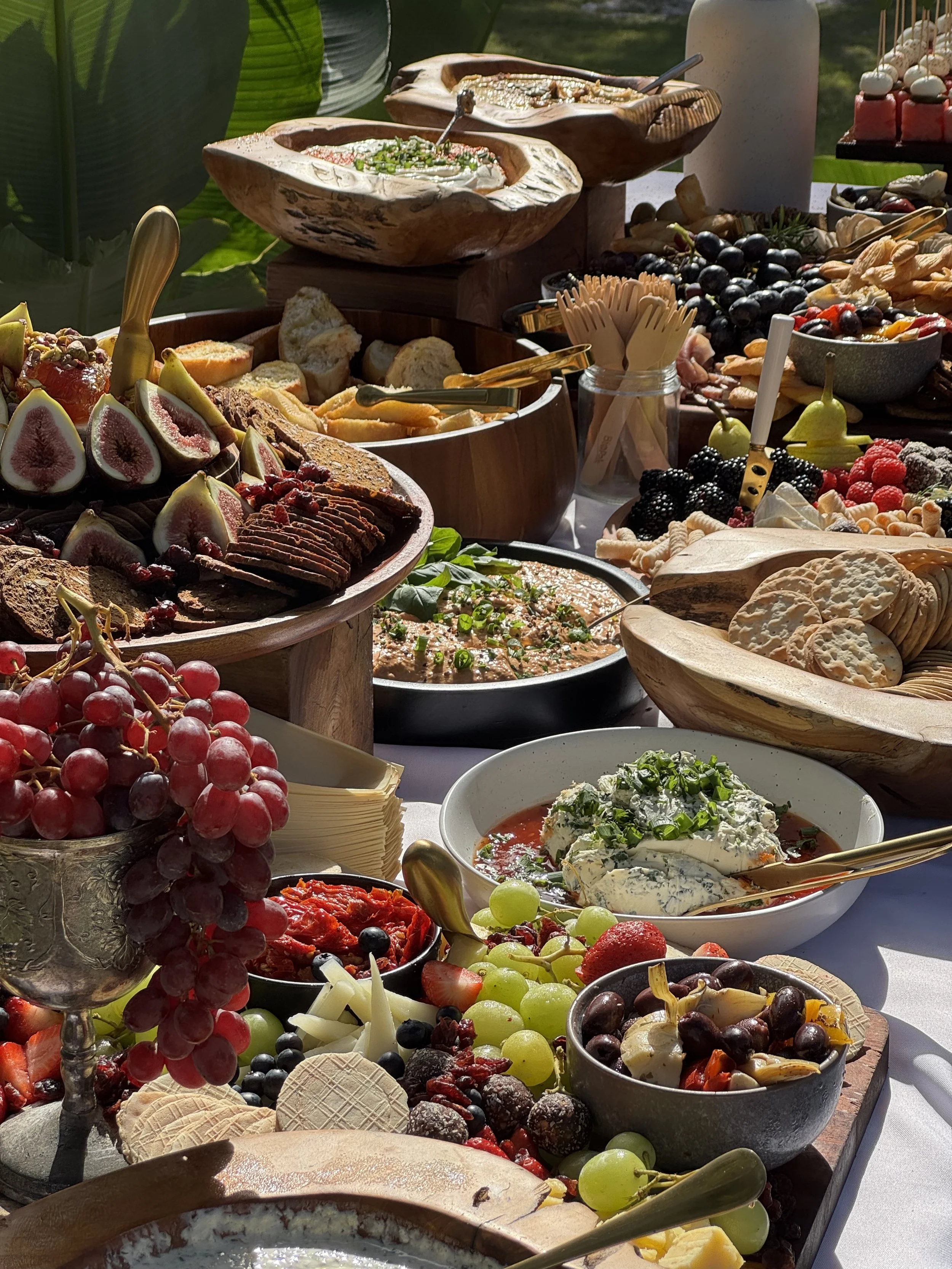 A variety of fresh fruit and cheese on a buffet table, including grapes, figs, berries, sliced bread, and a bowl of creamy cheese with herbs.