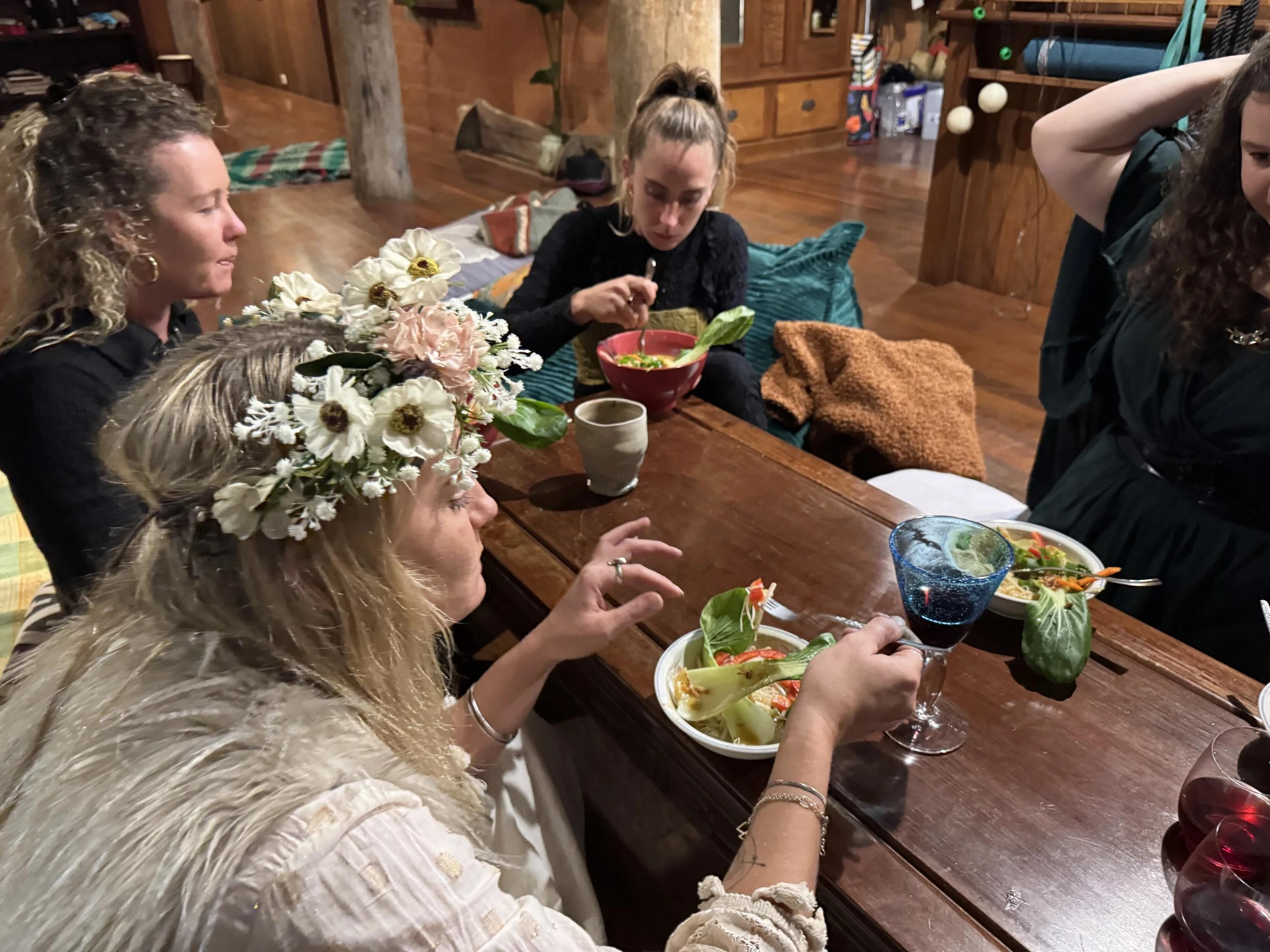People sitting around a wooden dining table, enjoying a meal in a cozy, rustic room with wood-paneled walls. There are flowers, some bowls of food, and drinks on the table.