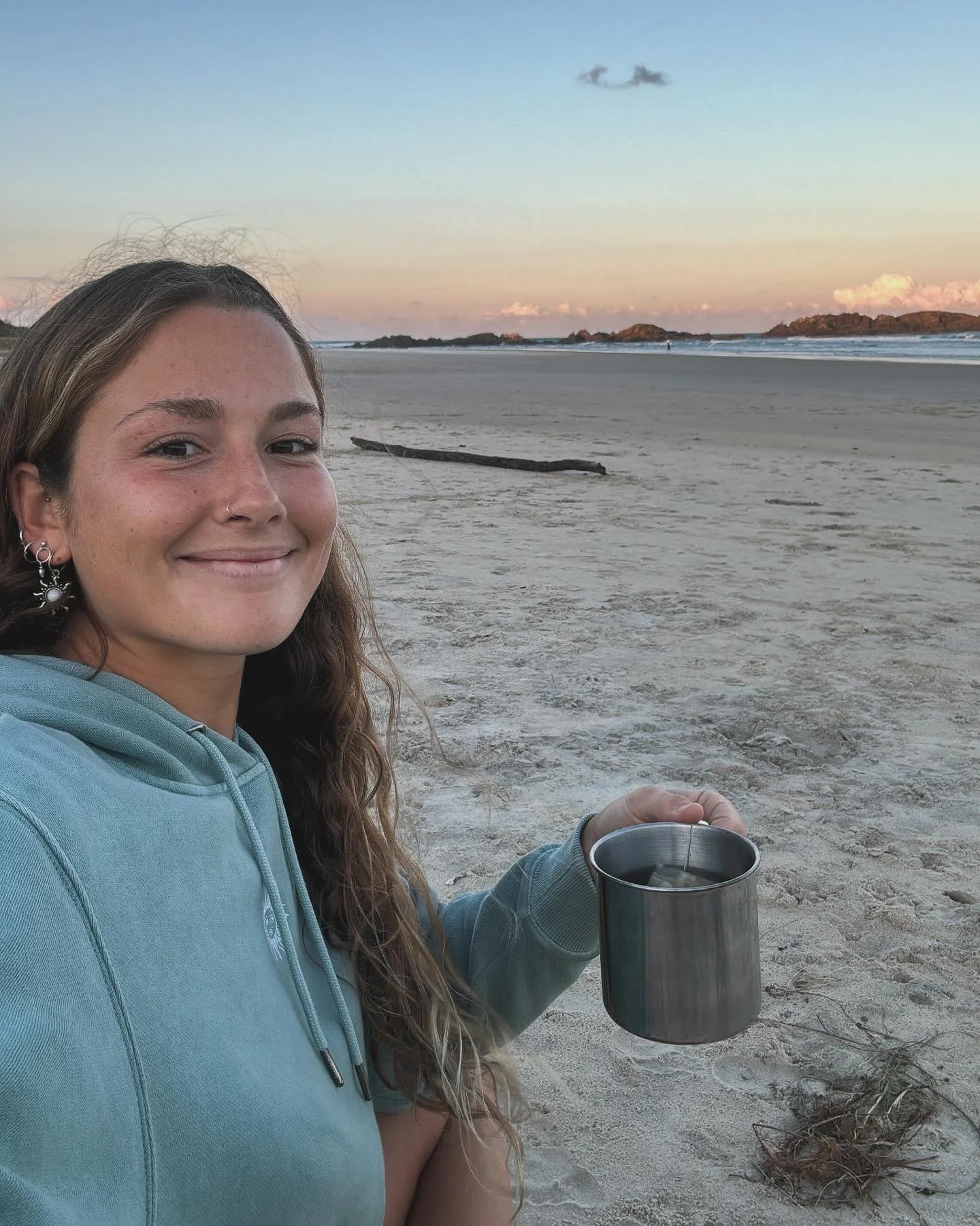 A young woman with long, brown hair, wearing a light blue hoodie, smiling while sitting on a sandy beach with a metal cup in her hand. The background features a calm ocean, rocky islands, and a colorful sunset sky with a few clouds.