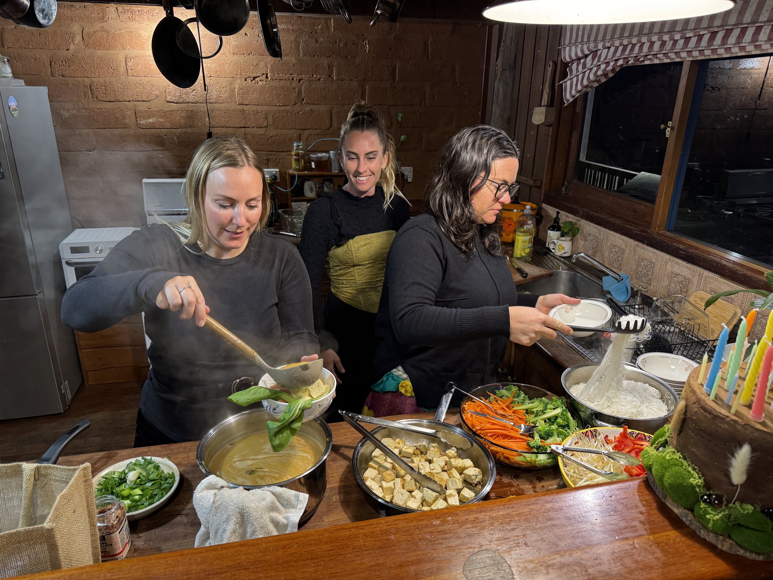 Three women in a kitchen preparing a meal, with various vegetables and dishes on the counter.