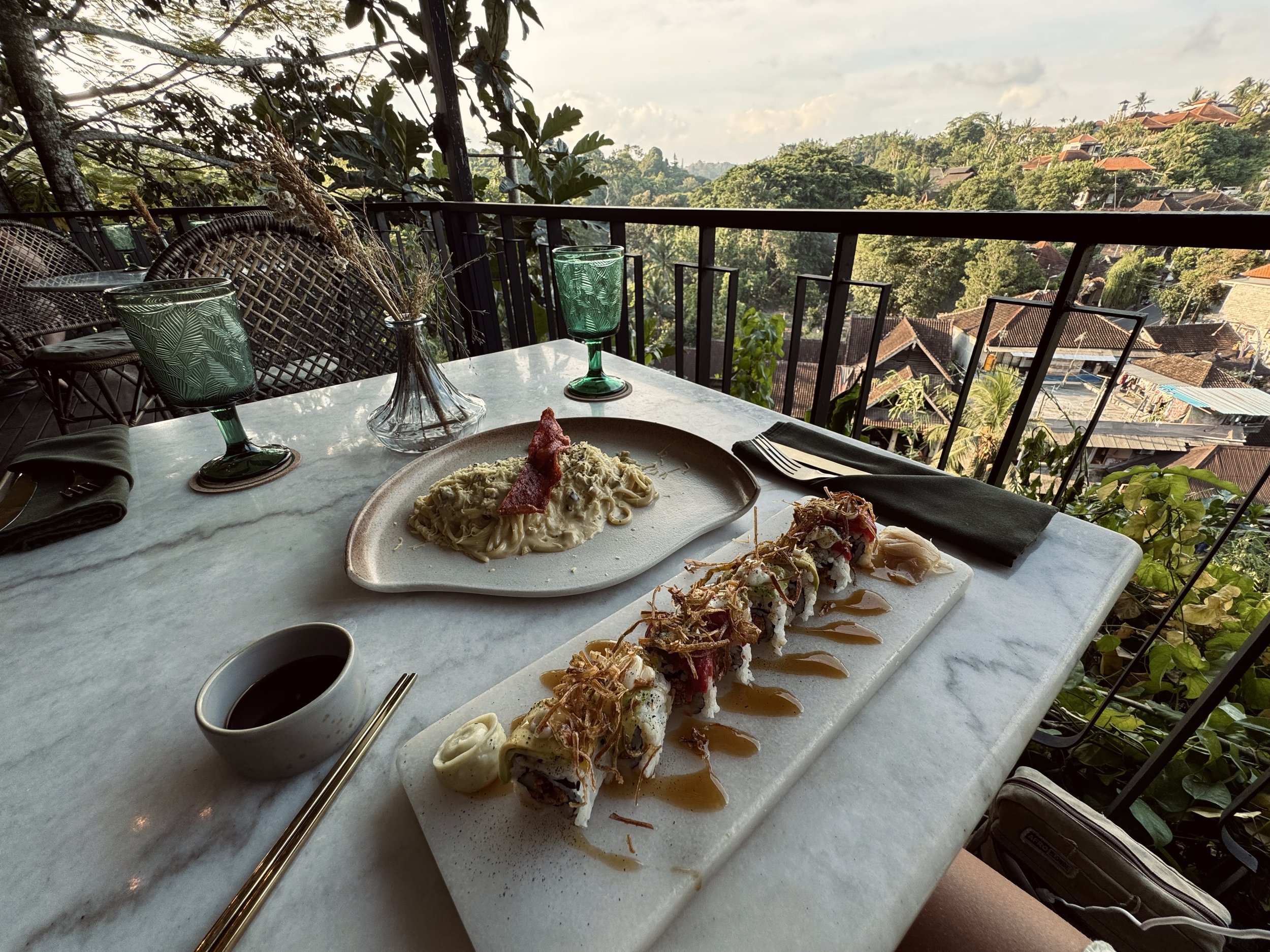 A table set with two green-tinted glasses, a small vase with dried flowers, a serving of pasta with sauce and bacon, and a small bowl of sauce. In the background, there is a balcony railing overlooking a lush, green hillside with houses and trees.